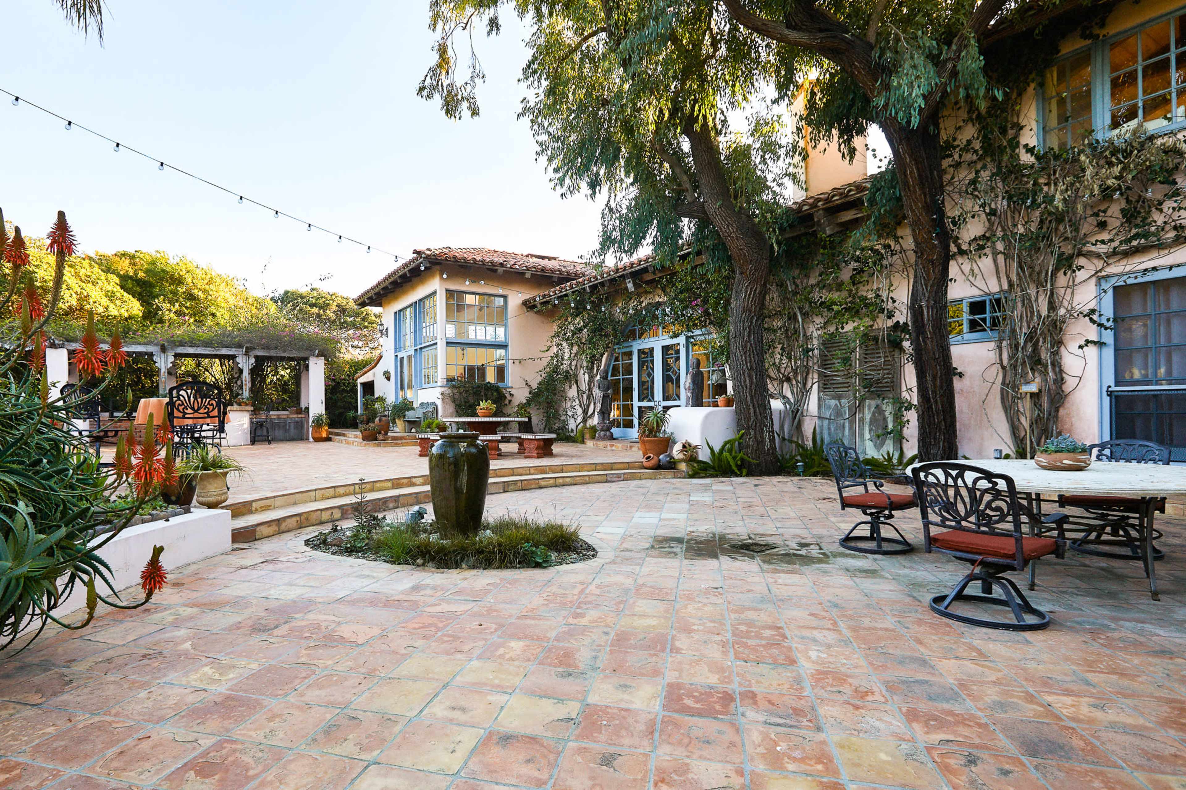 A spacious outdoor courtyard surrounded by a mix of buildings, featuring tiled flooring, potted plants, and outdoor seating.