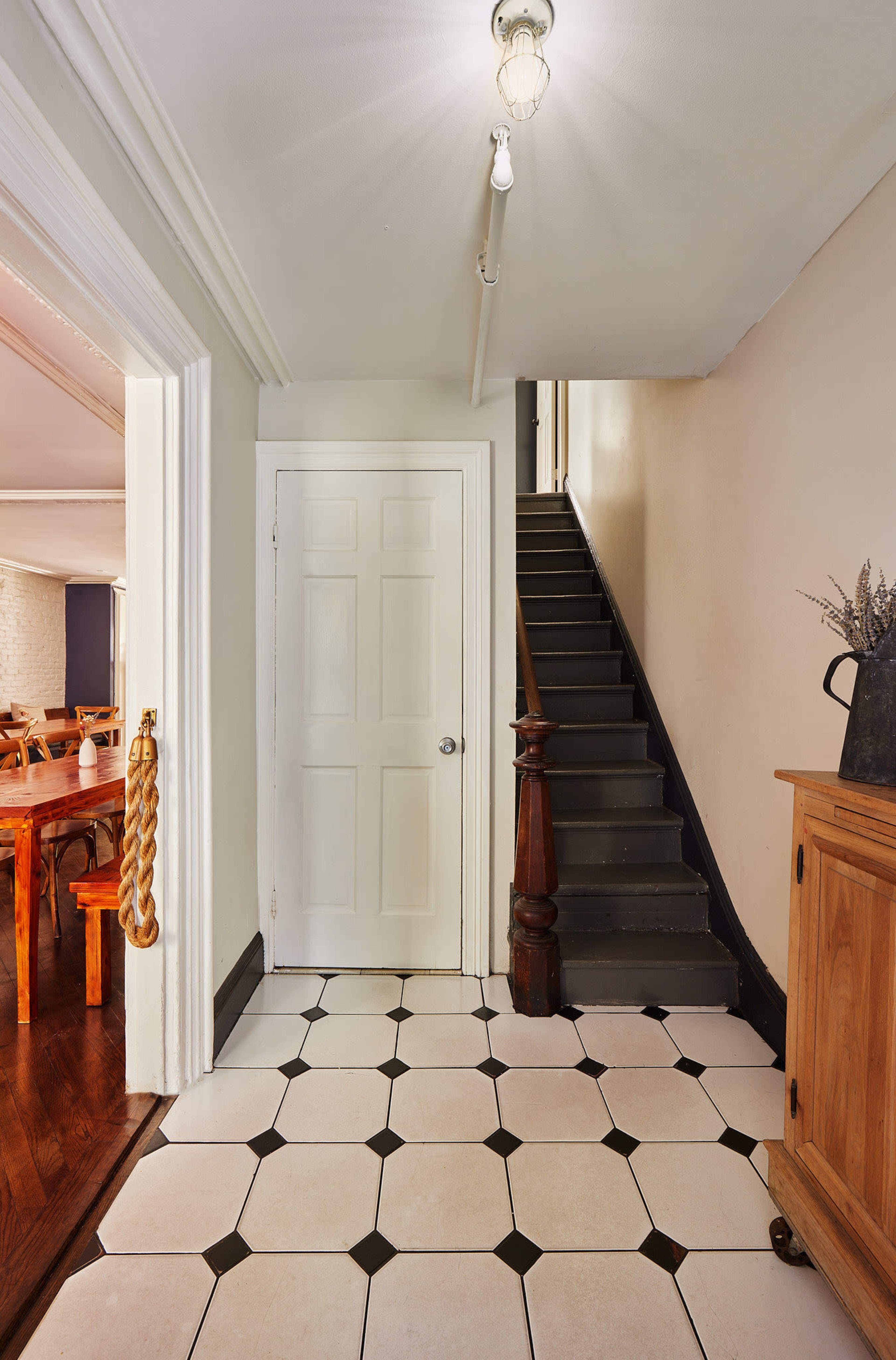 The image shows a hallway with black and white tiled flooring, a closed door on the left, a staircase leading upward, and a wooden sideboard on the right.