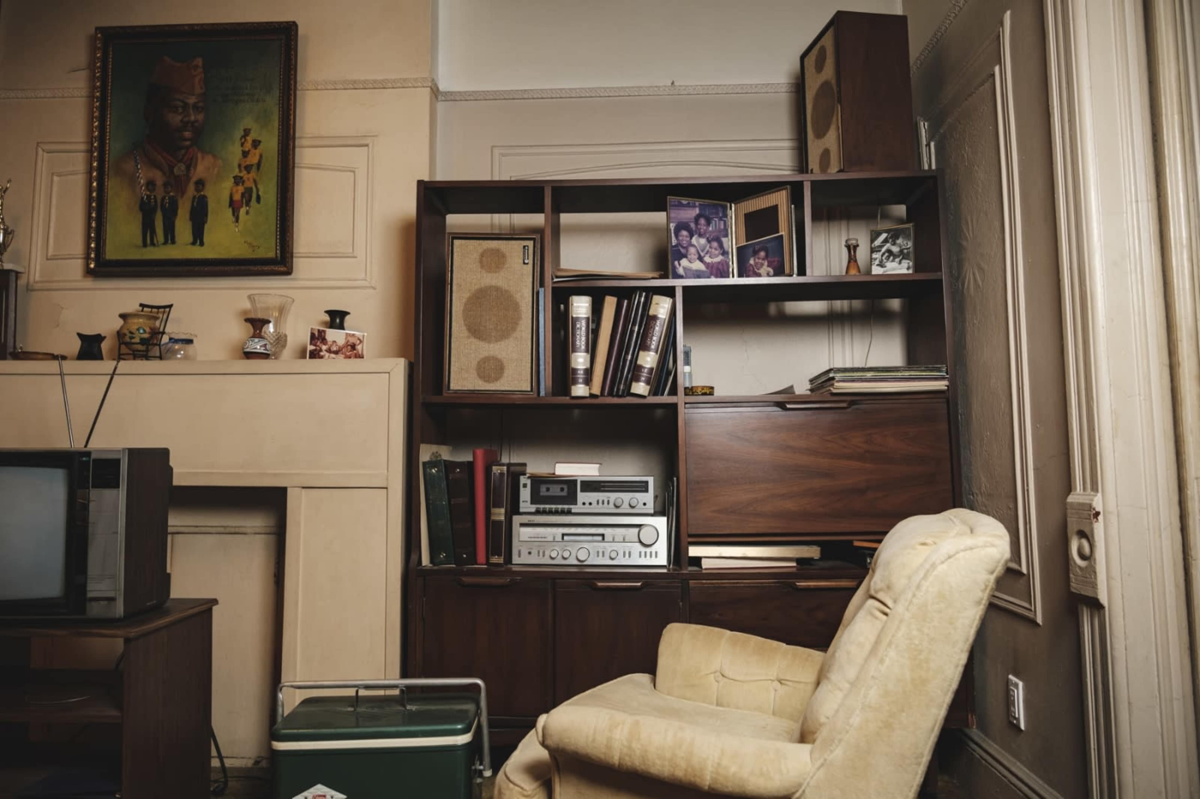 A corner of a room featuring a wooden shelf with books, photographs, and audio equipment, alongside a vintage television and a light-colored armchair.