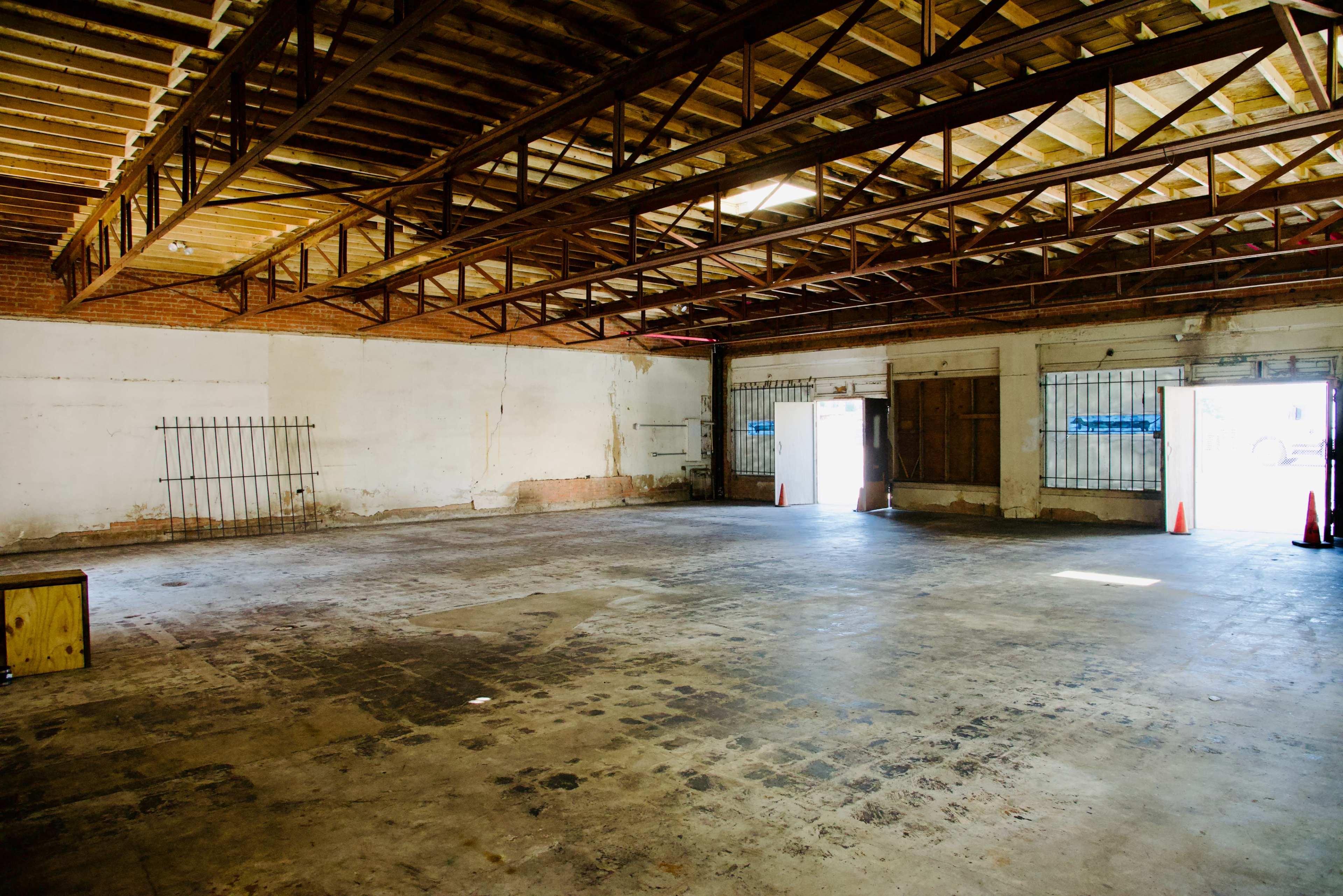 An empty warehouse interior with exposed wooden beams, concrete flooring, and two open doors leading outside.