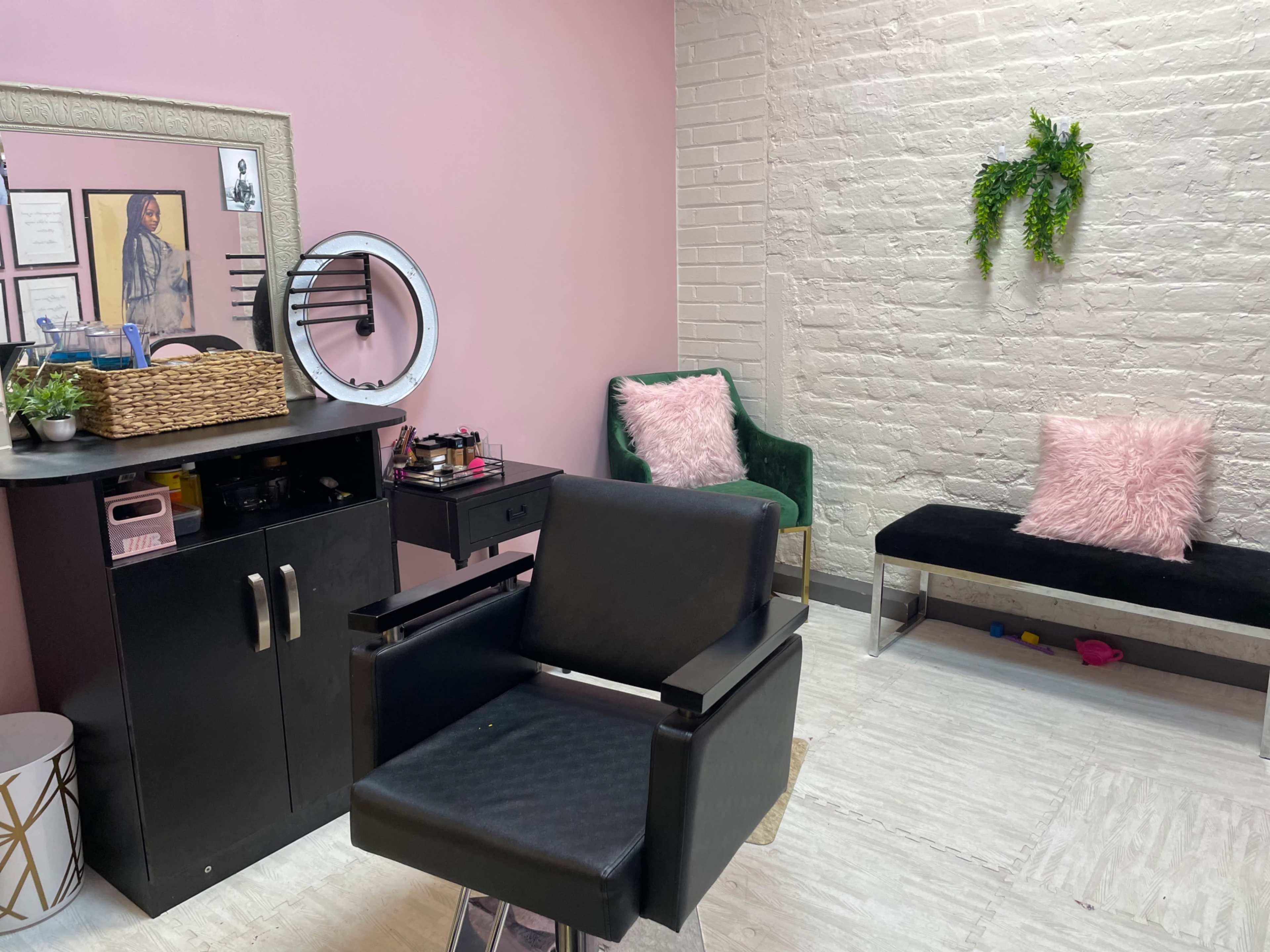 A hair salon interior with a styling chair, a small black workstation, a pink accent wall, and two green chairs with a decorative plant on the wall.