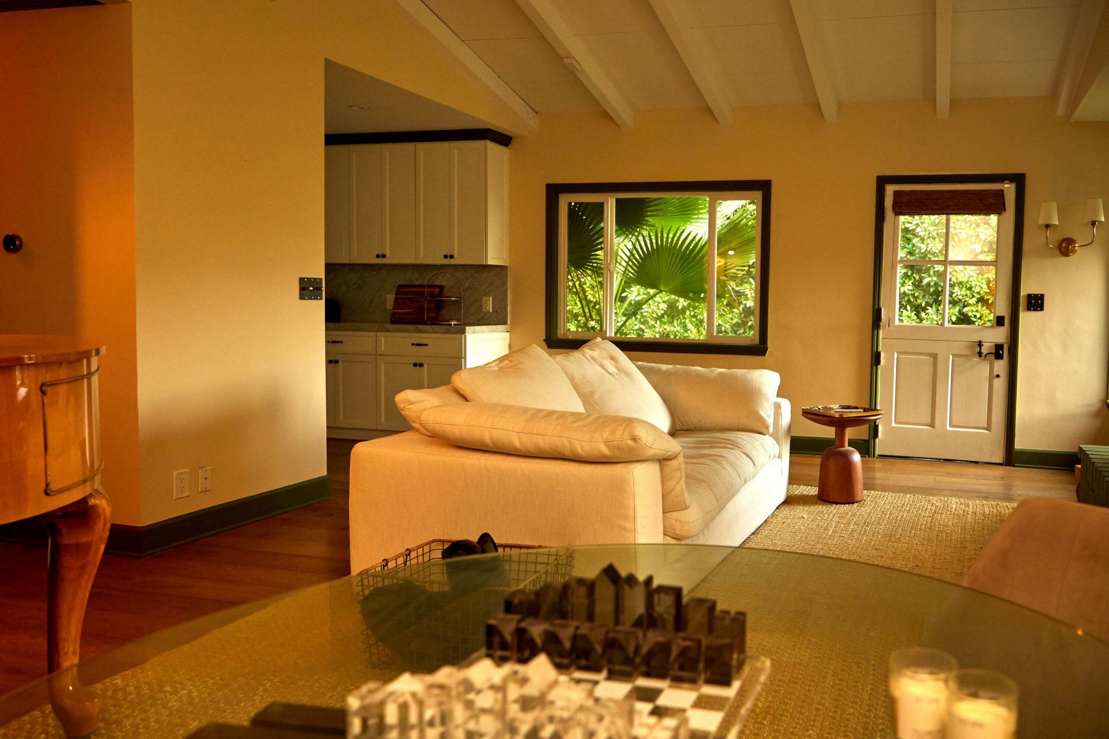 The image shows a cozy living room with a large white sofa, a coffee table displaying a chess set, and a view of greenery through windows.