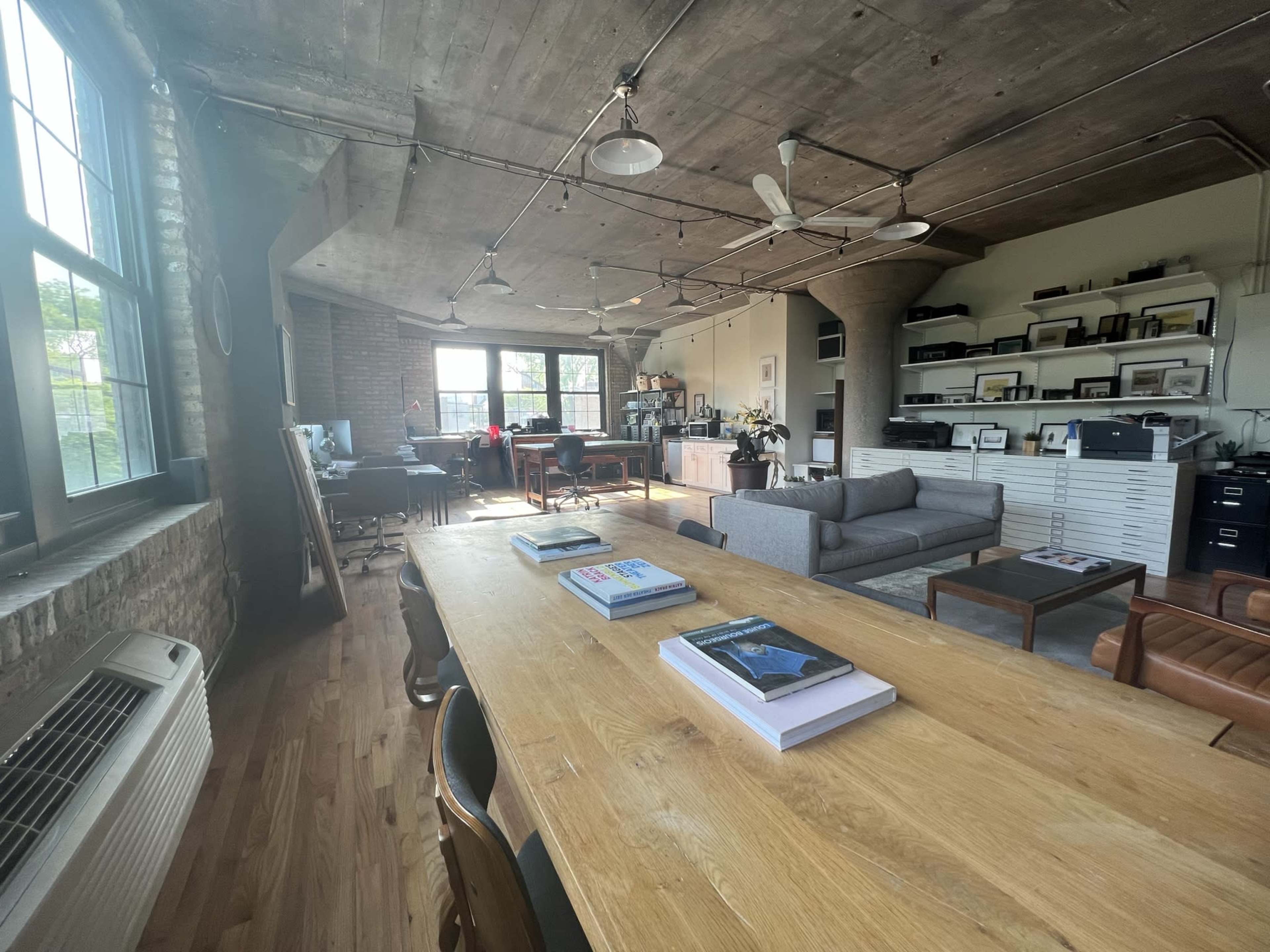 A spacious, well-lit office with a large wooden table, a gray couch, and shelves filled with various items against exposed brick walls and industrial-style ceilings.