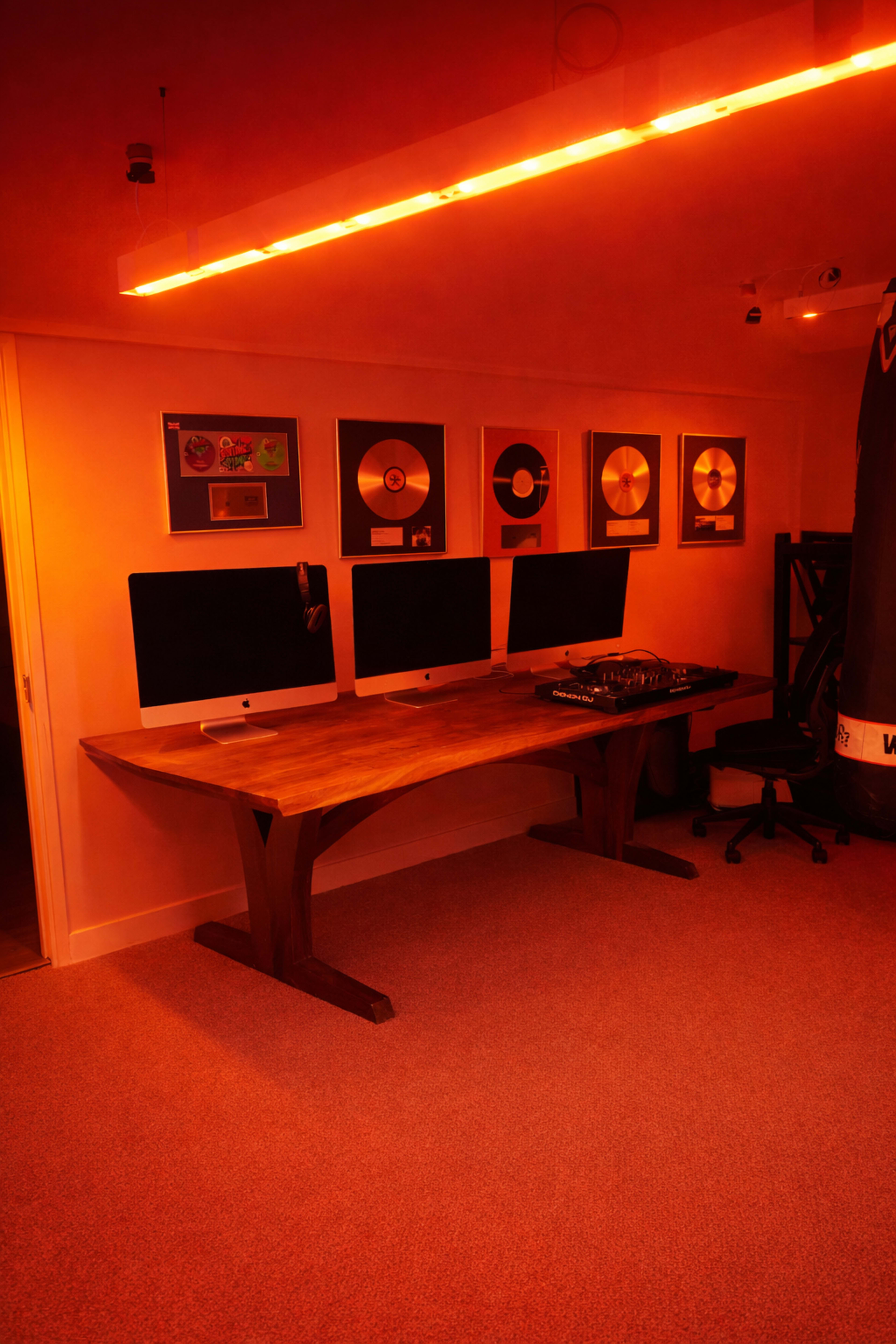 The image shows a modern workspace with three Apple monitors on a wooden desk, illuminated by orange lighting and featuring framed awards on the wall.
