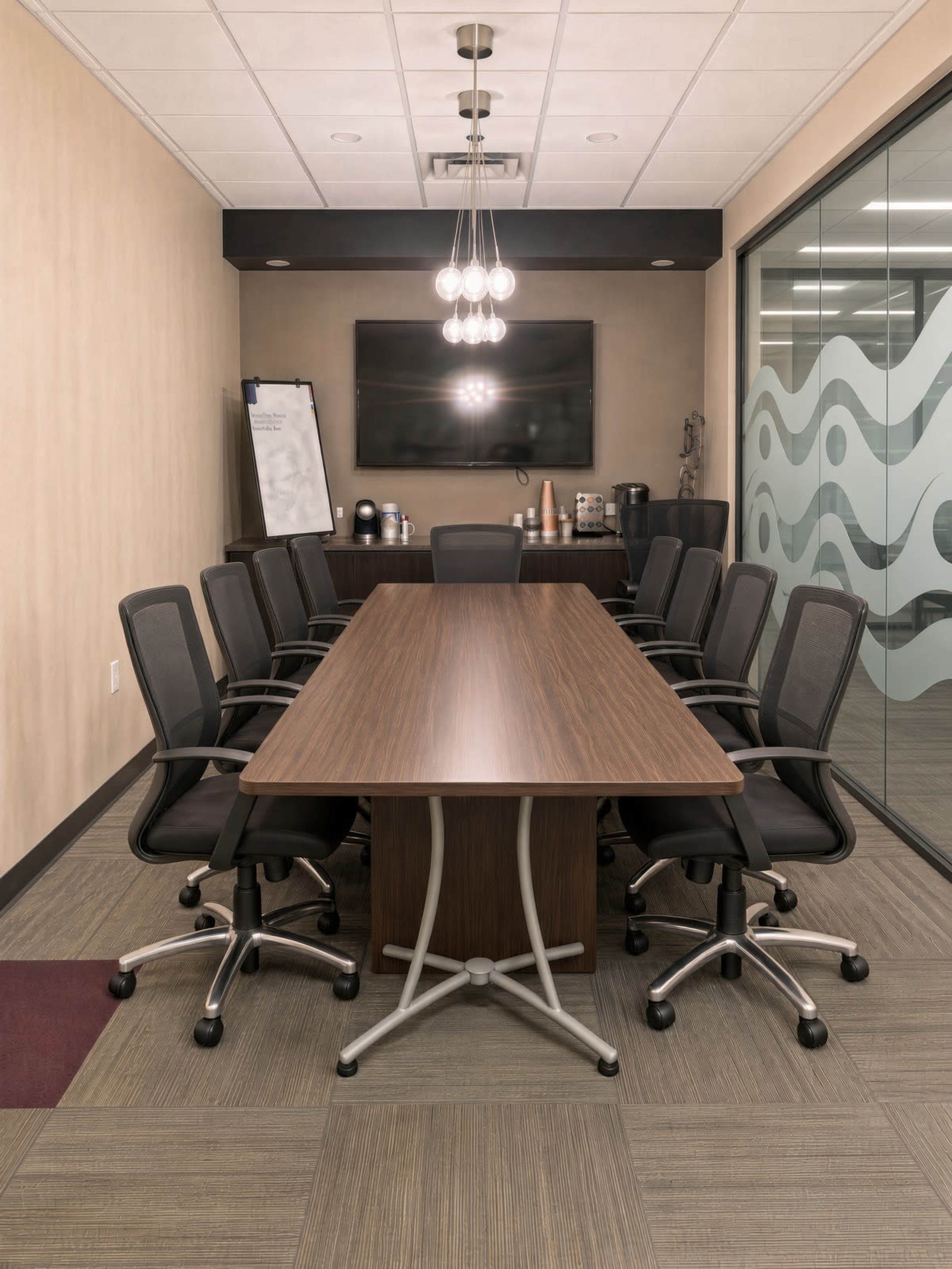 A modern conference room features a long wooden table surrounded by ergonomic office chairs, with a large screen and coffee station in the background.
