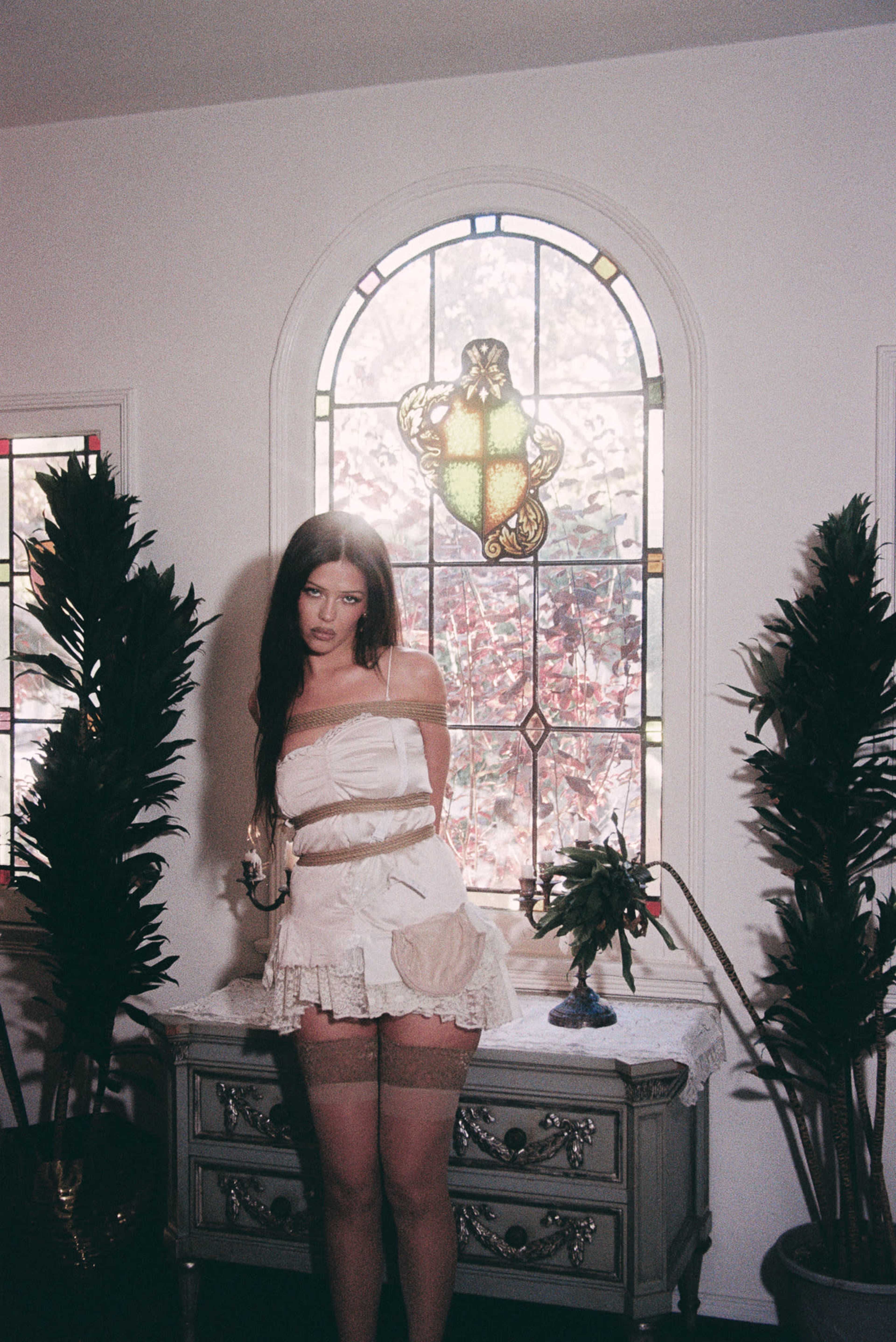 A woman stands in a room with a stained glass window, flanked by potted plants and beside a vintage dresser.