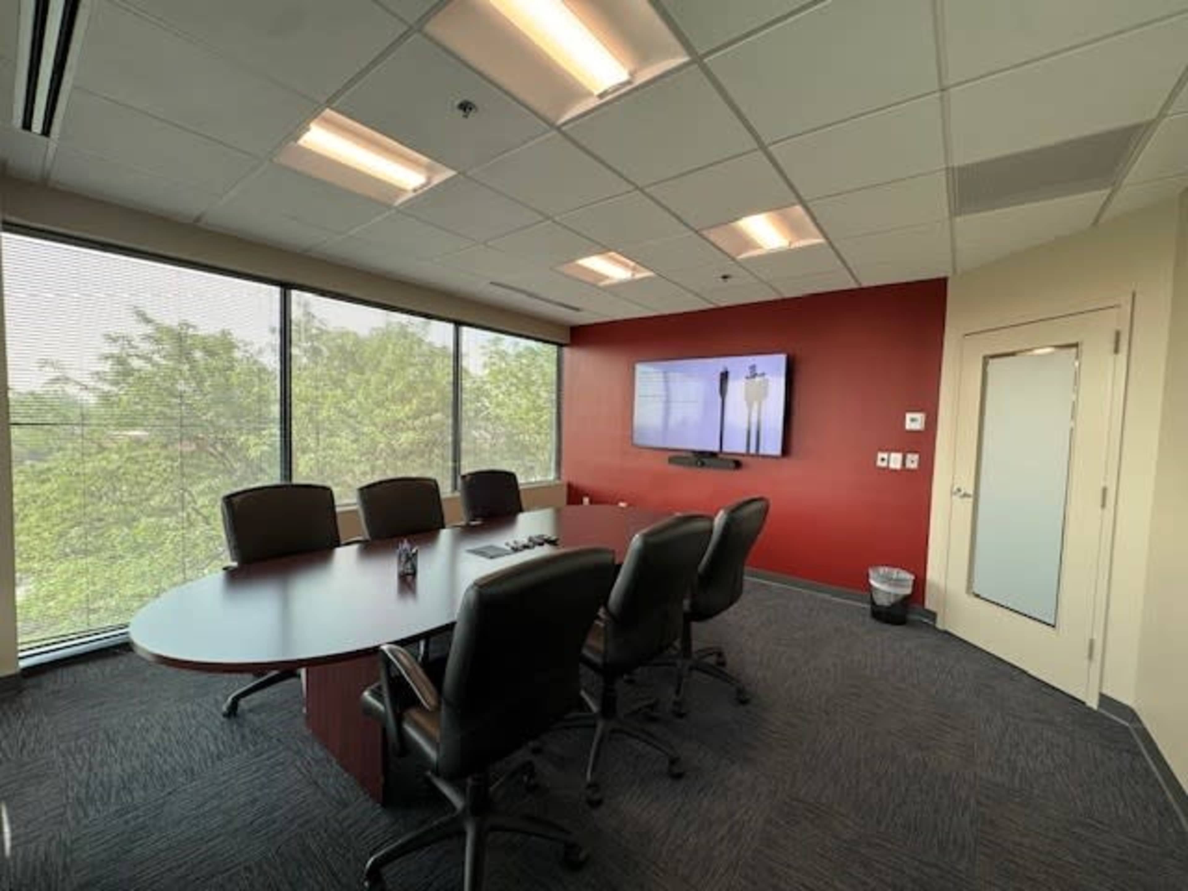 The image shows a conference room with a large oval table, several black chairs, and a wall-mounted screen, surrounded by windows that provide natural light.