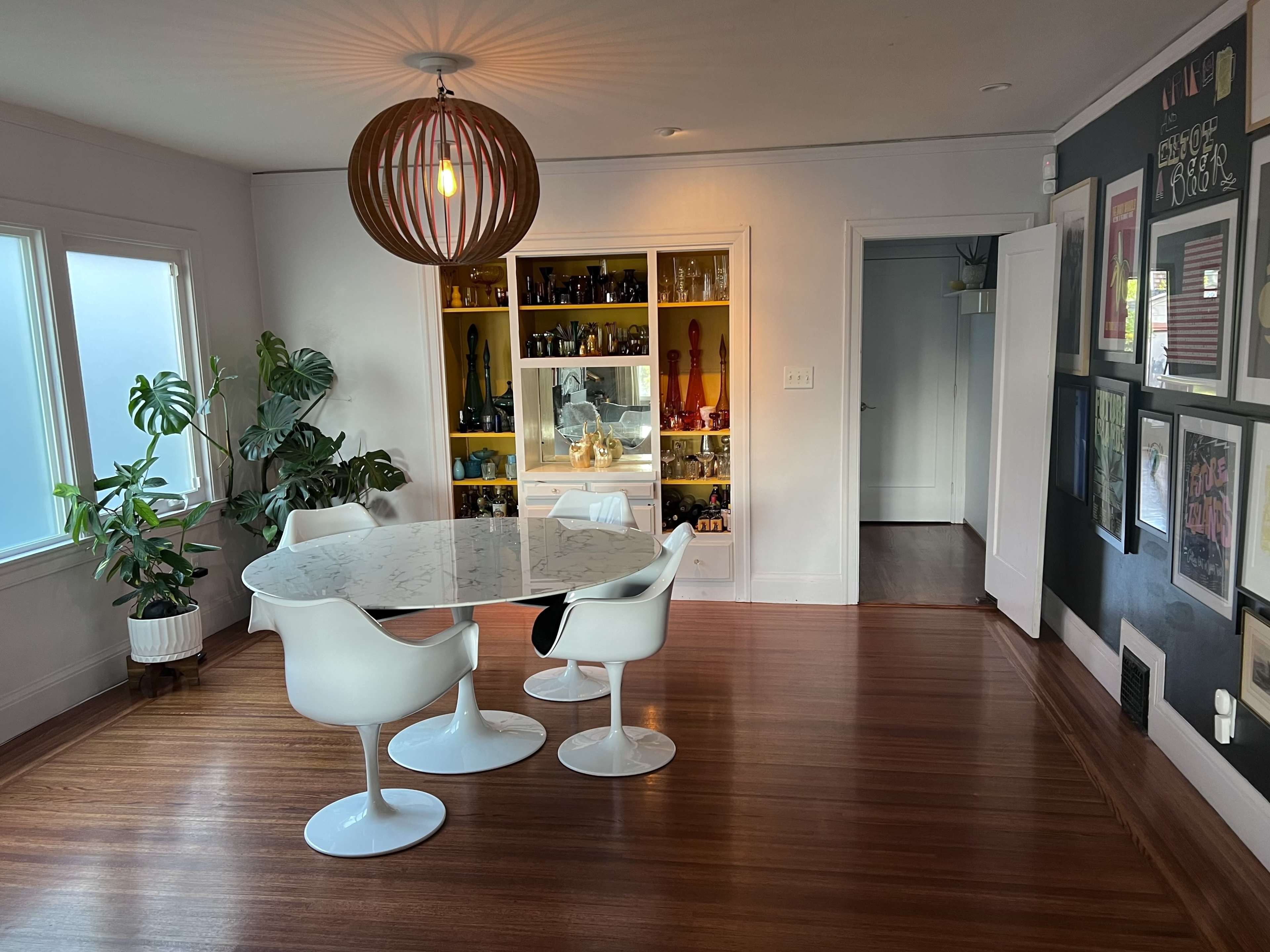 A modern dining area features a round marble table with white chairs, illuminated by a pendant light, with a cabinet displaying various items against a wall with framed pictures.