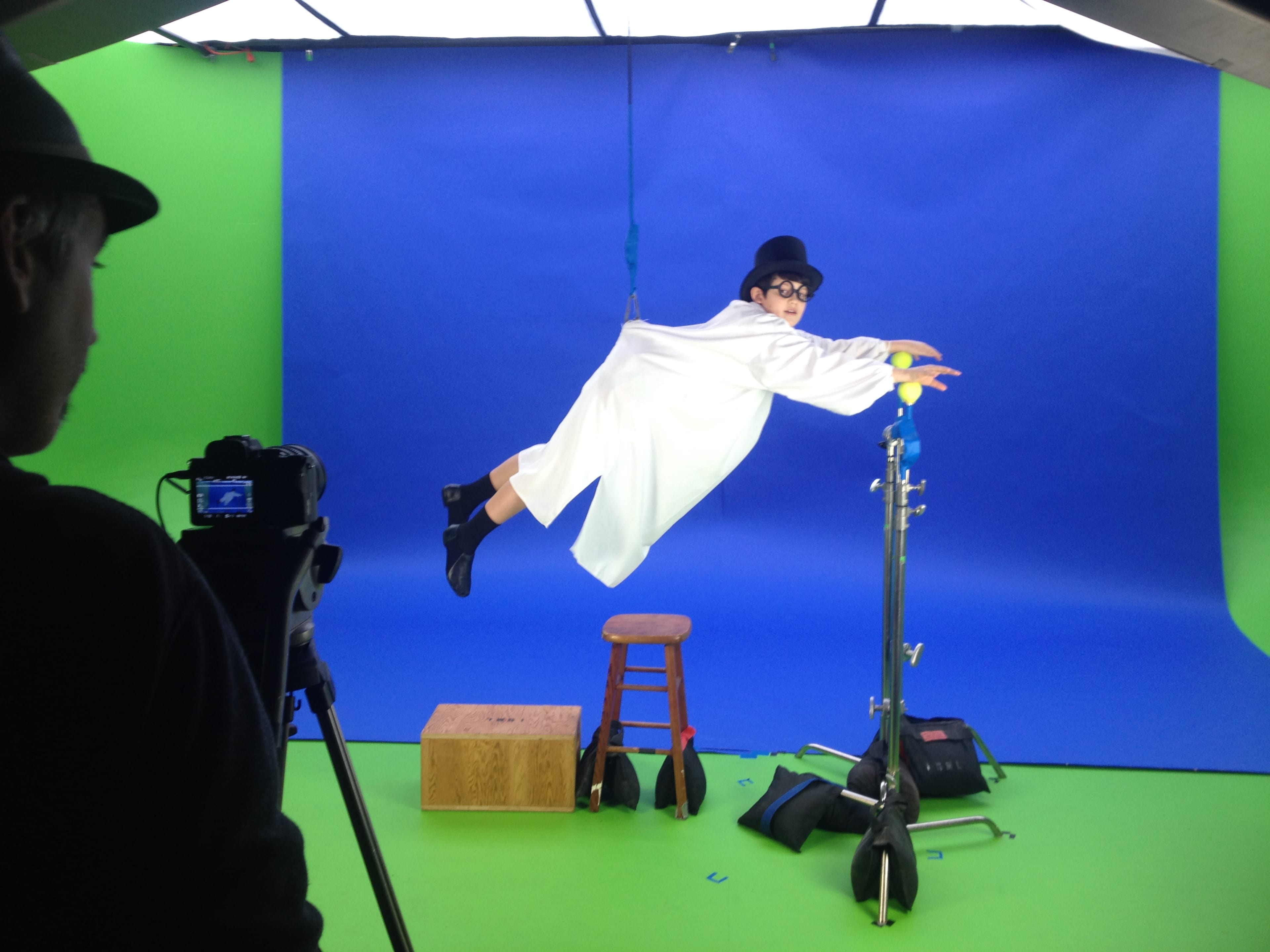 A person in a white coat and bowler hat is suspended in mid-air against a blue and green backdrop, while another person operates a camera nearby.