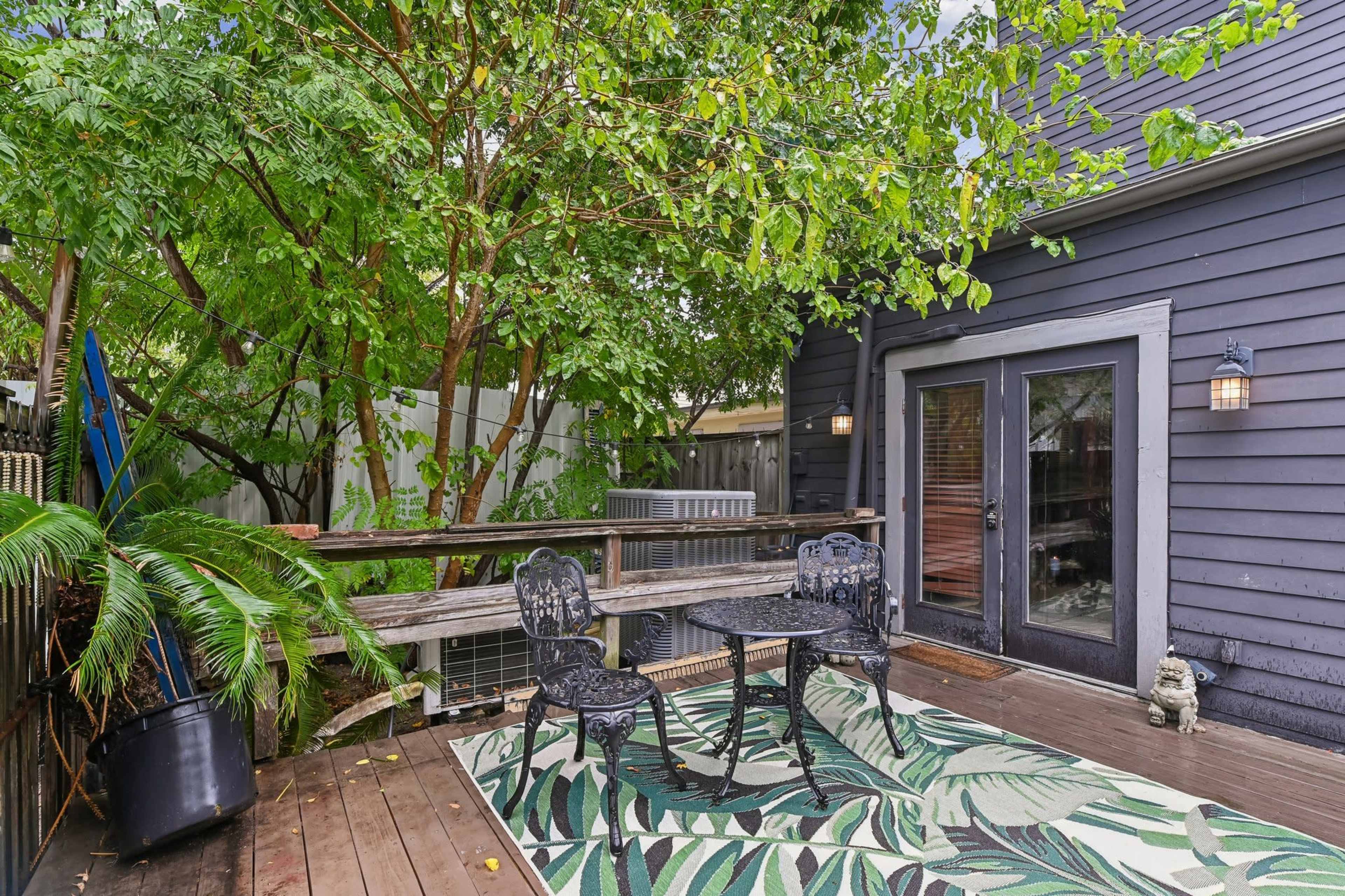 A small wooden deck features a table and two chairs, surrounded by lush greenery and a decorative outdoor rug.