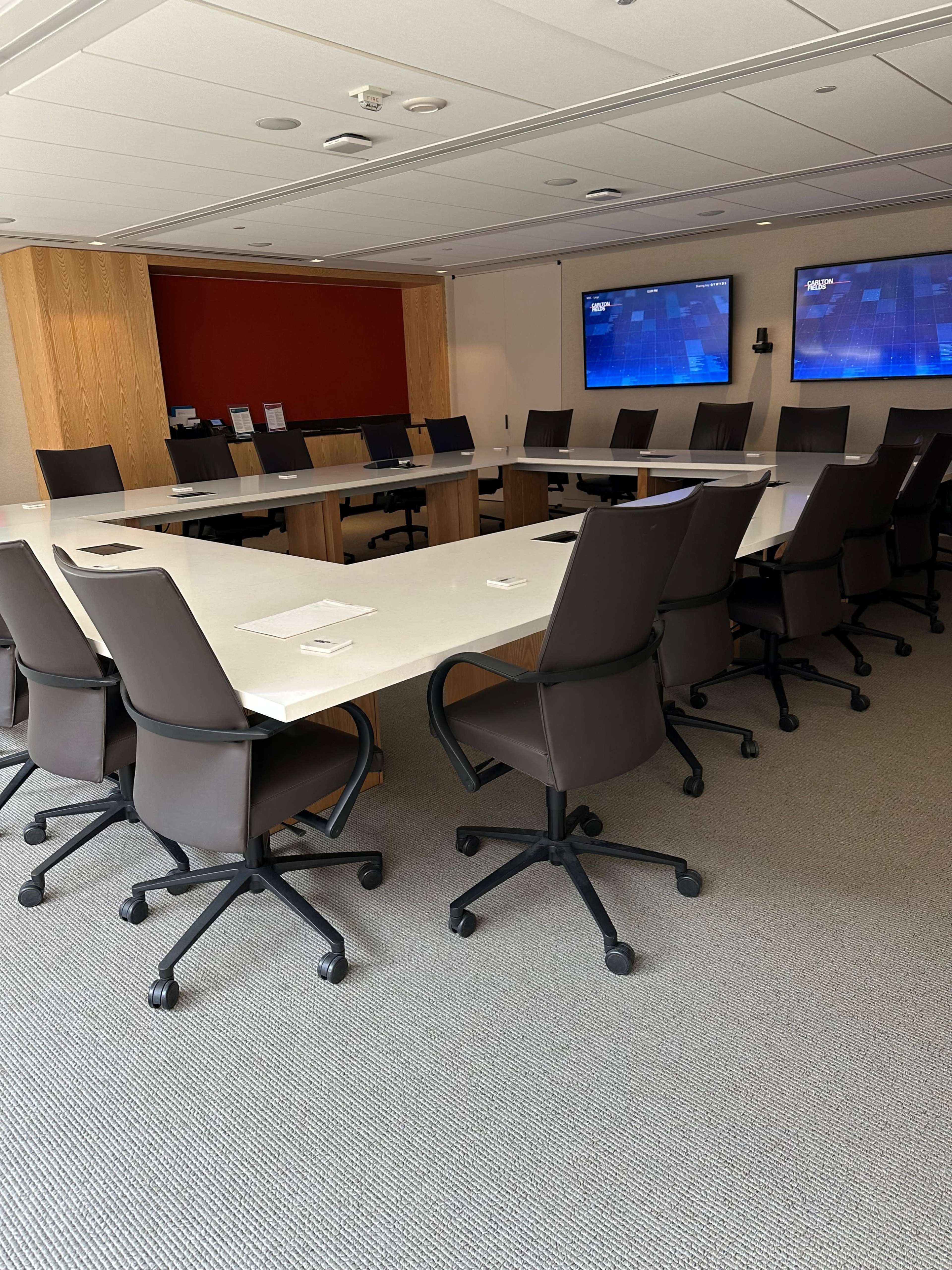 A large conference room features a rectangular table surrounded by ergonomic chairs, with two screens mounted on the walls.