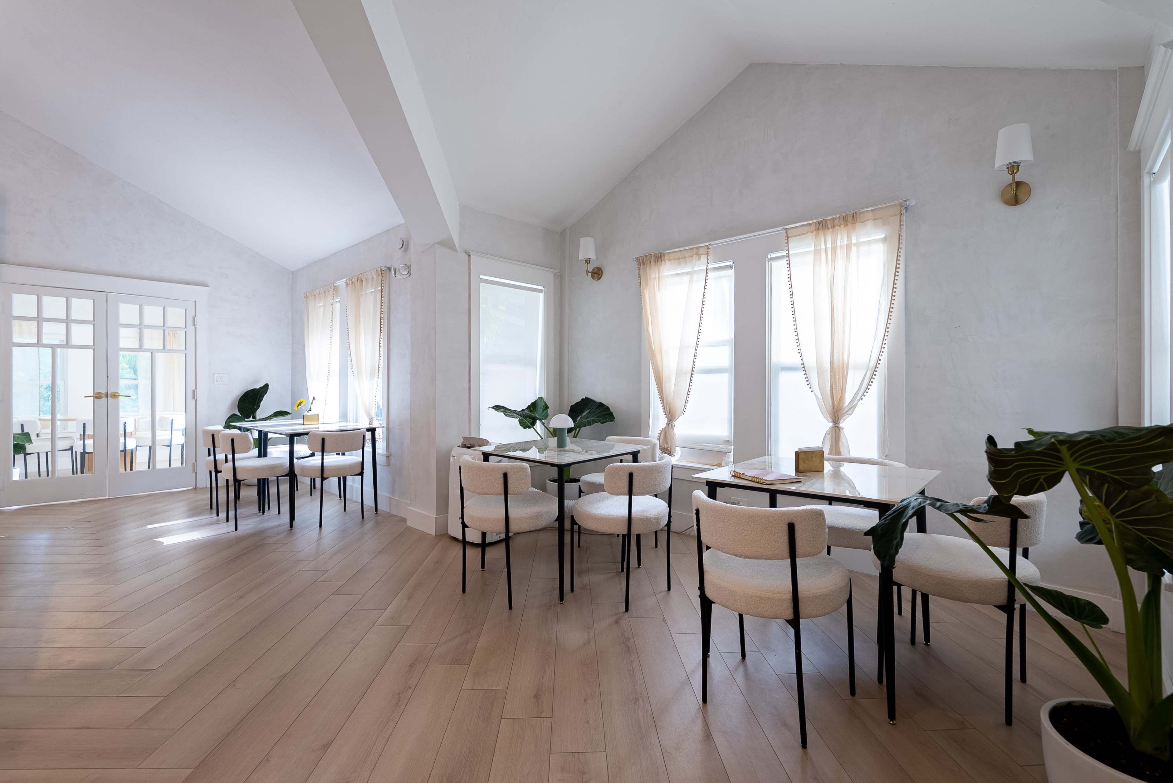 The image shows a bright, modern café interior with wooden flooring, several tables, and large windows draped with light-colored curtains.