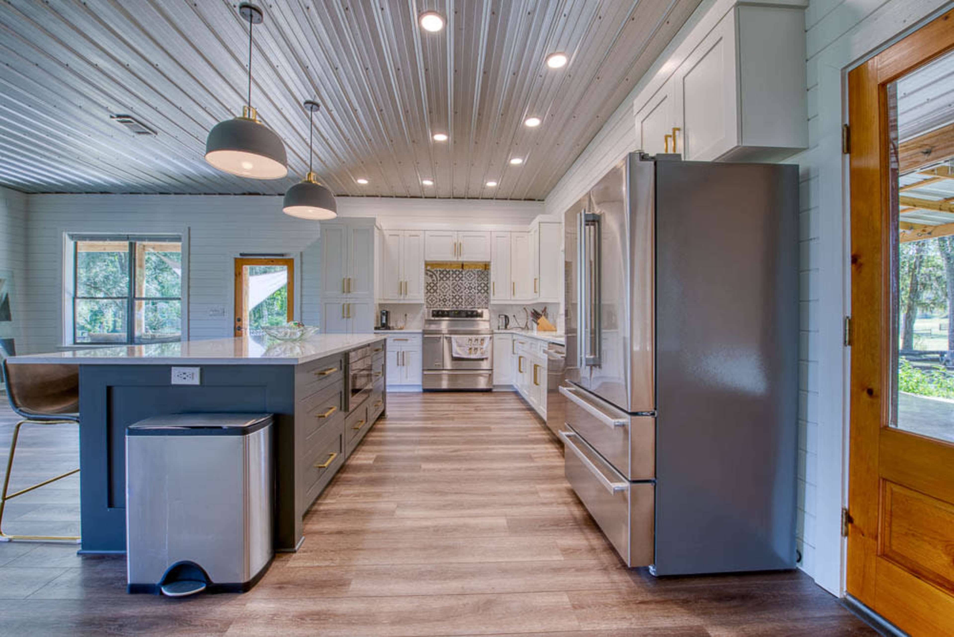 A modern kitchen features a large island, stainless steel appliances, and white cabinetry with natural light streaming through the windows.