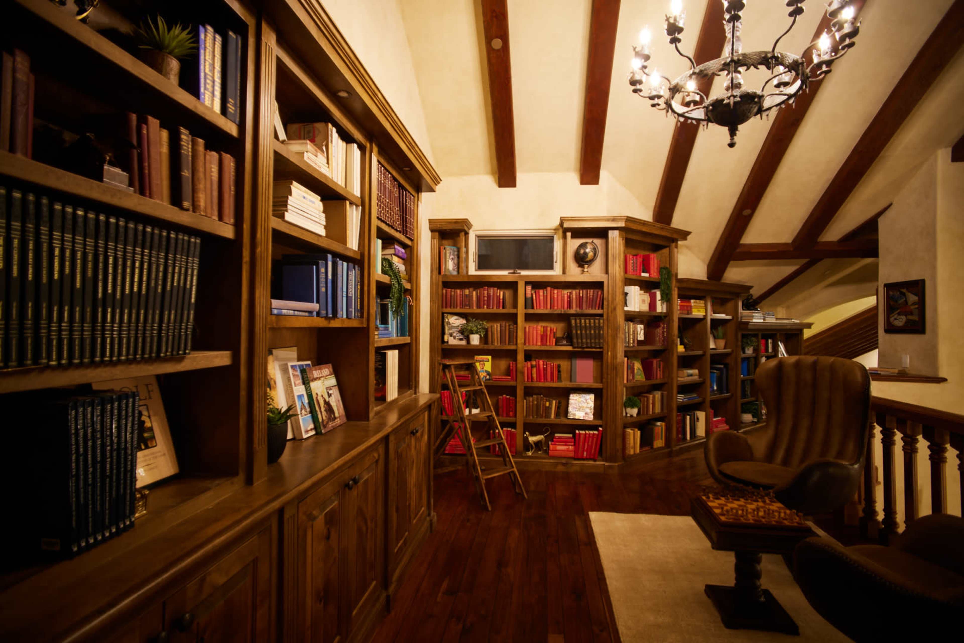 A warmly lit library with wooden bookshelves filled with books, a ladder, and a plush armchair beneath a decorative chandelier.