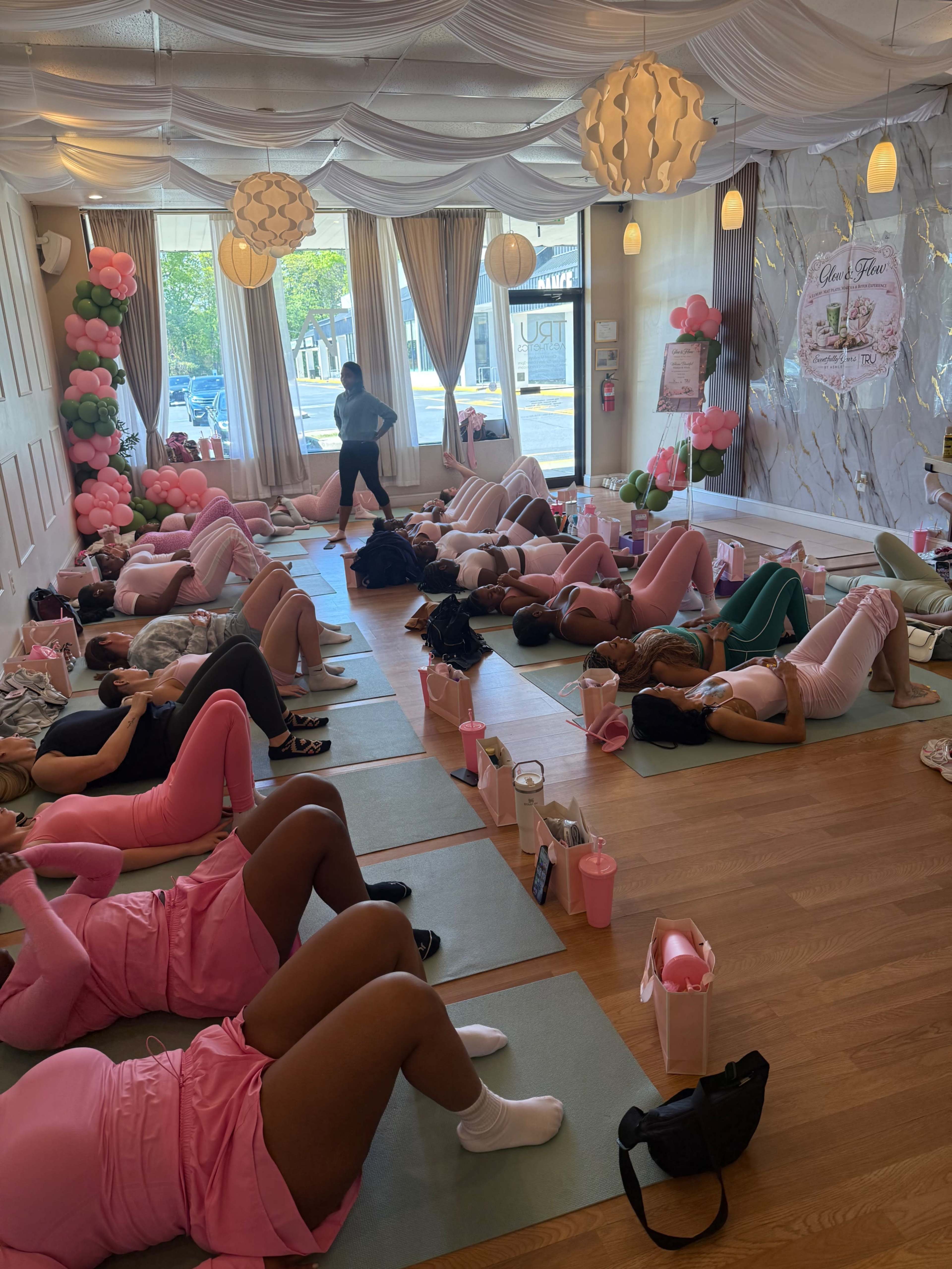 A group of women in pink attire lies on yoga mats in a bright studio decorated with balloons and soft lighting, participating in a fitness class.