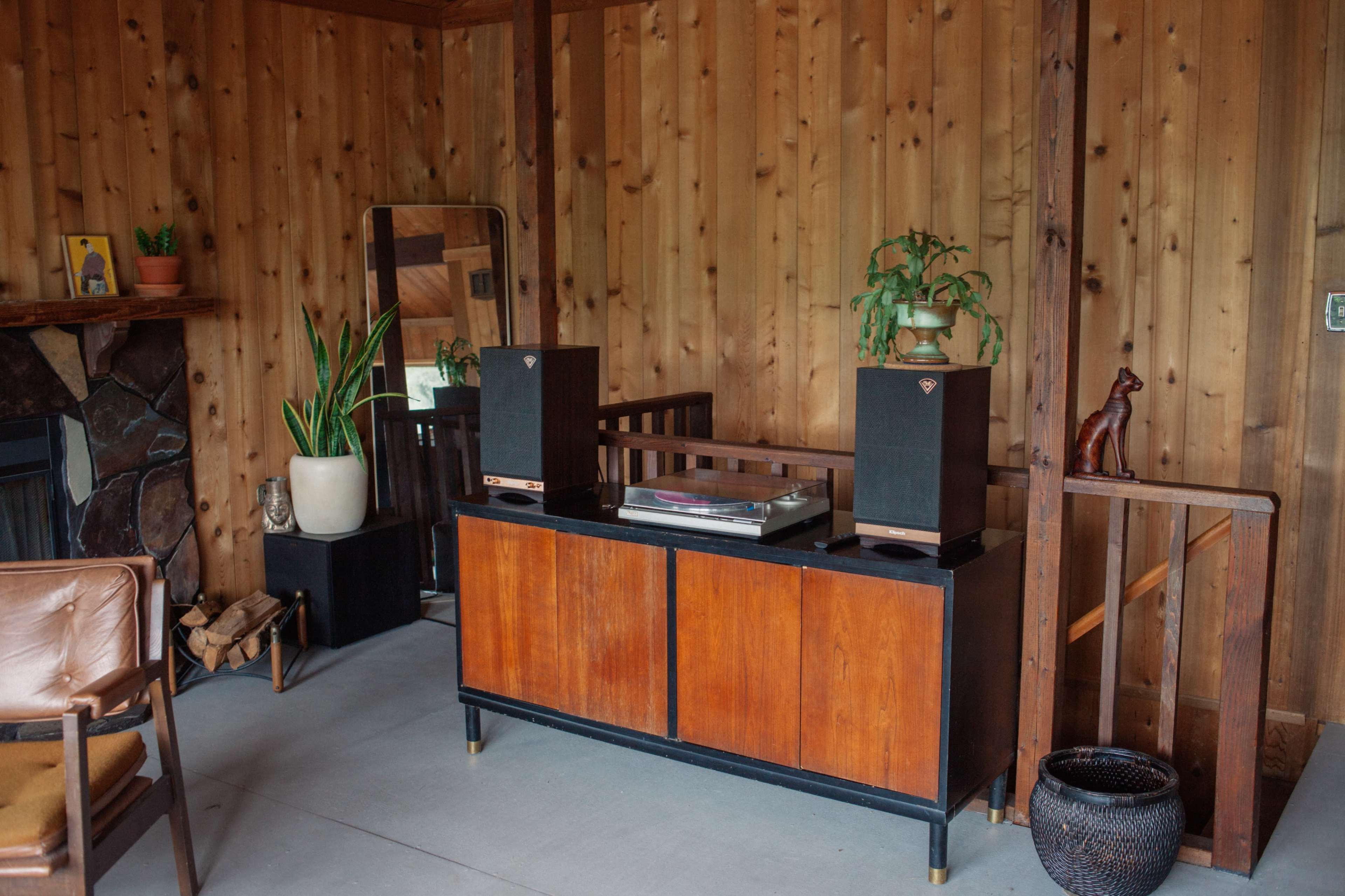 A wooden room with a console holding speakers and a turntable, surrounded by plants and a mirror, with a vintage chair nearby.