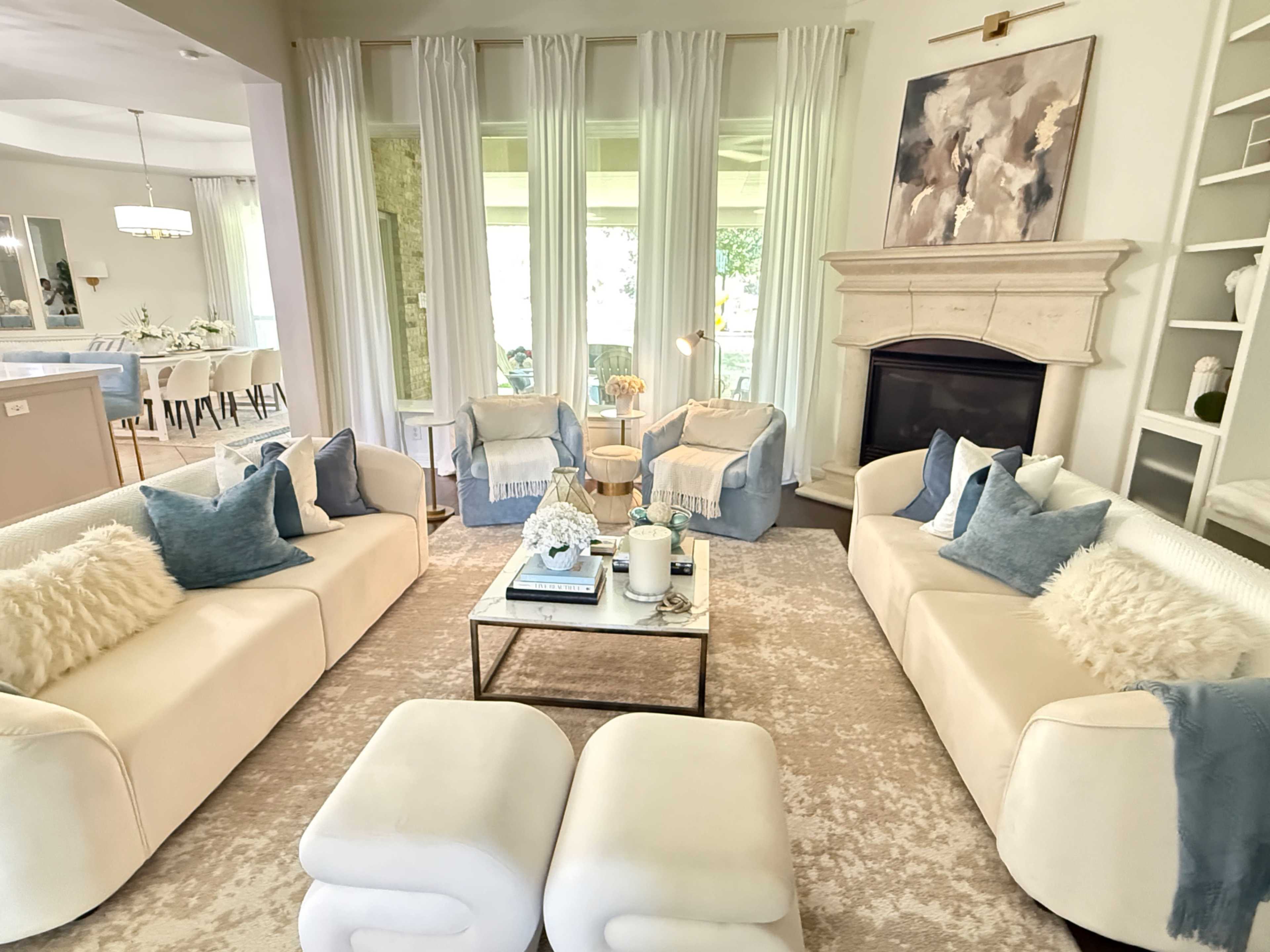 The living room features two cream-colored sofas facing each other, accompanied by light blue armchairs, a coffee table in the center, and a fireplace framed by large windows.