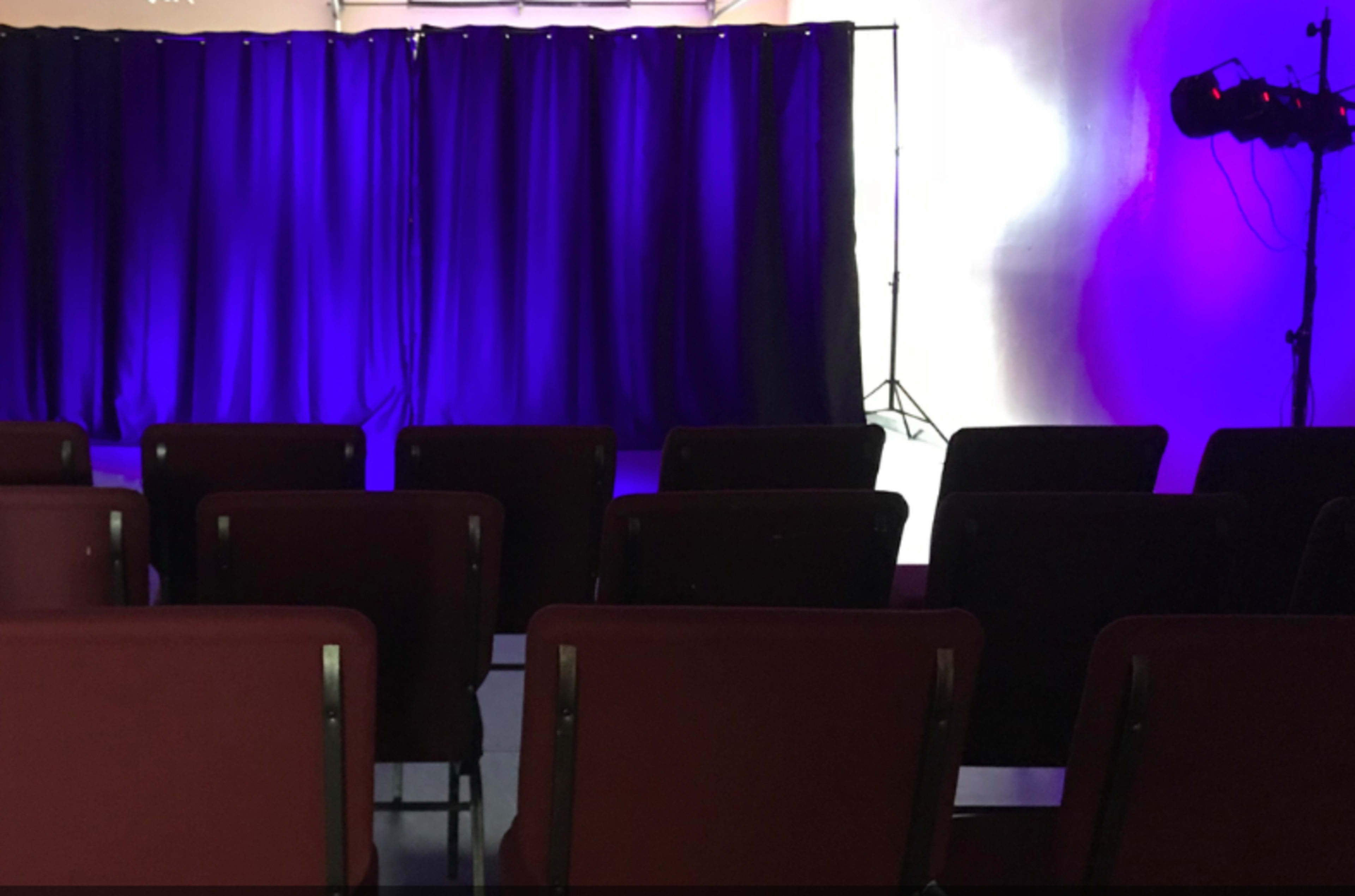 A row of red chairs facing a stage with a backdrop of purple curtains and lighting.