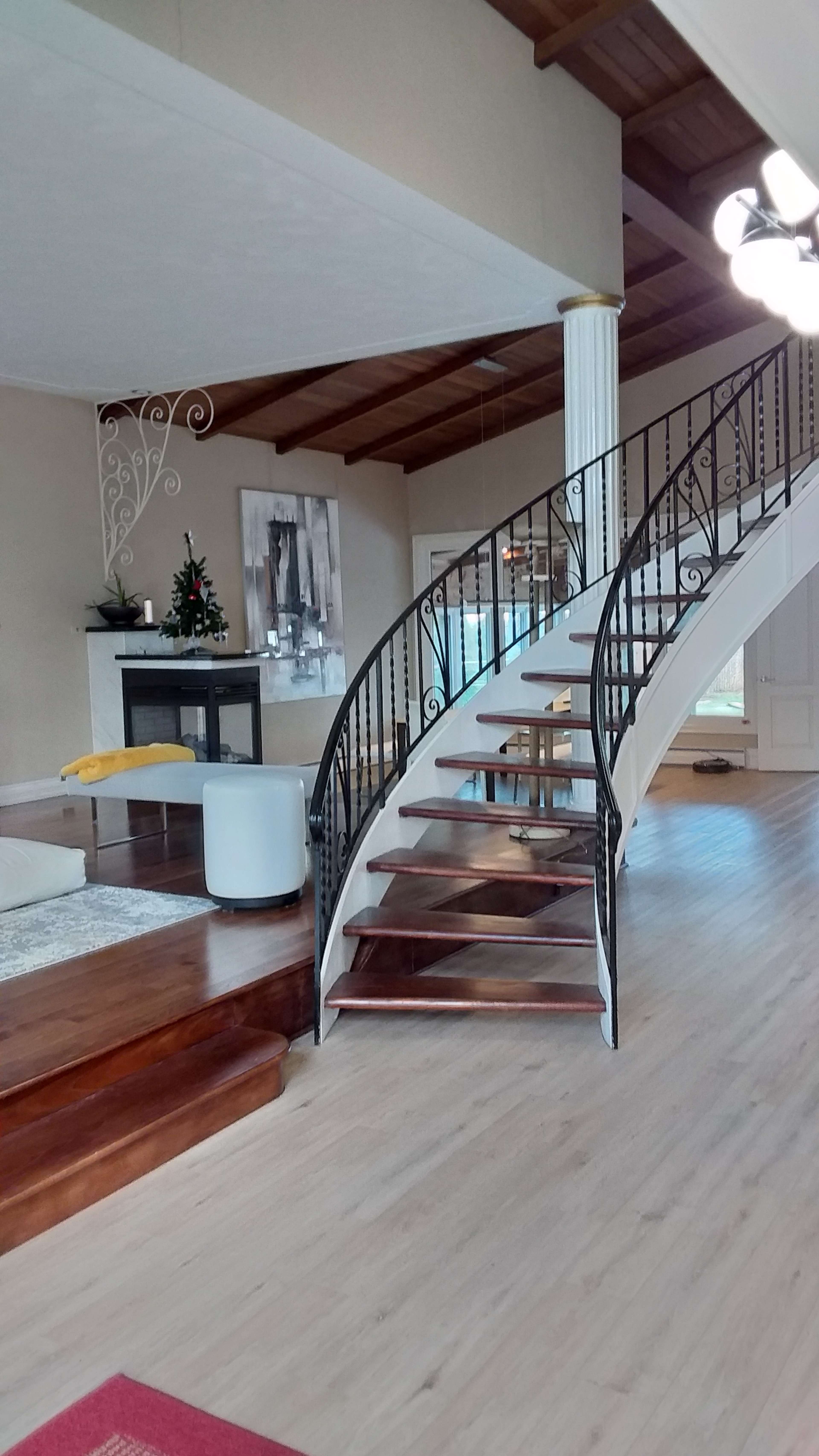 A spacious living area with a staircase featuring wooden steps and an ornate black railing, leading to an upper level, alongside a modern fireplace and decorative elements on the wall.