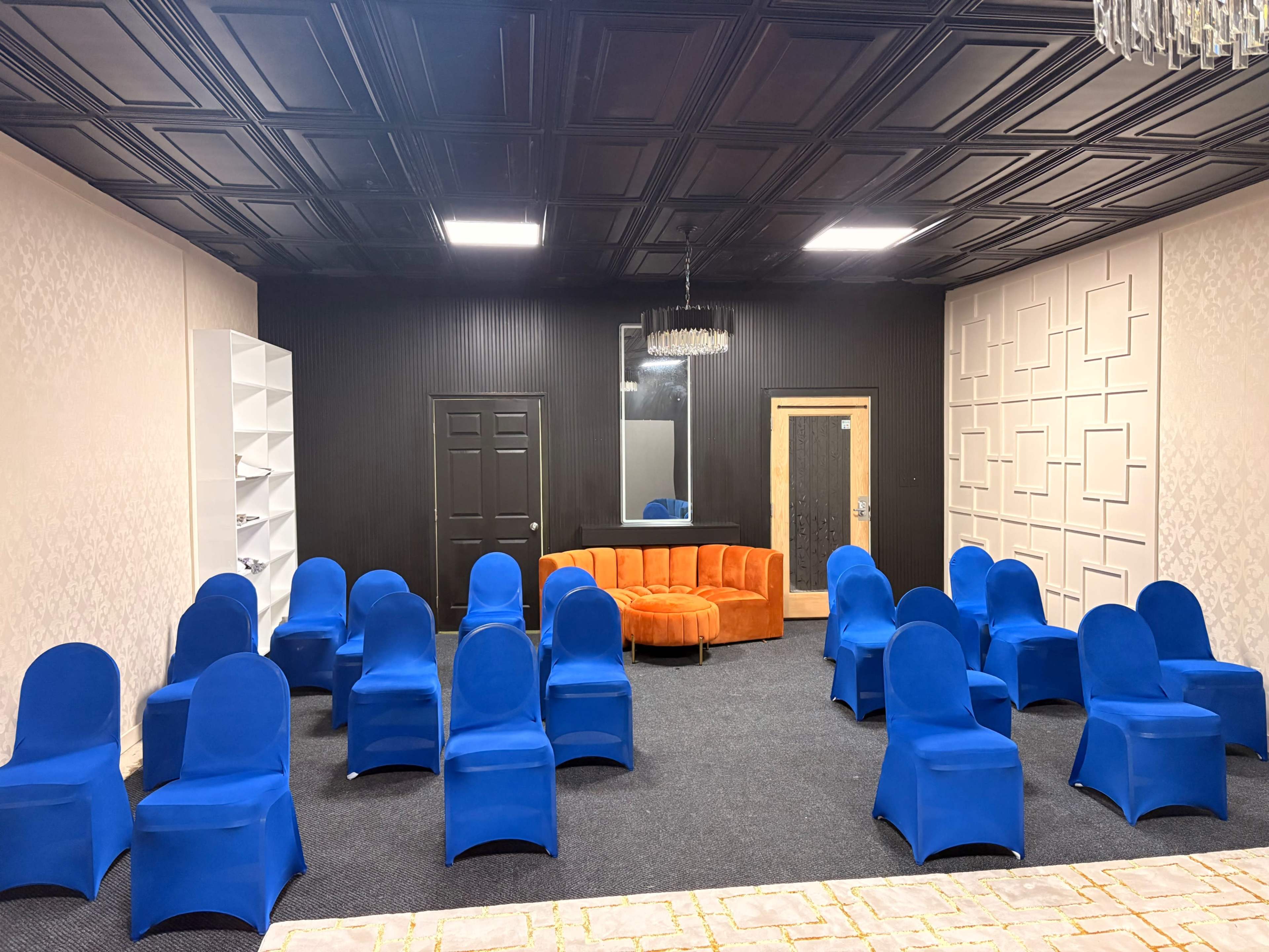 The image shows a room with an orange circular couch and neatly arranged blue chairs facing a black wall, with a decorative ceiling and light fixtures.