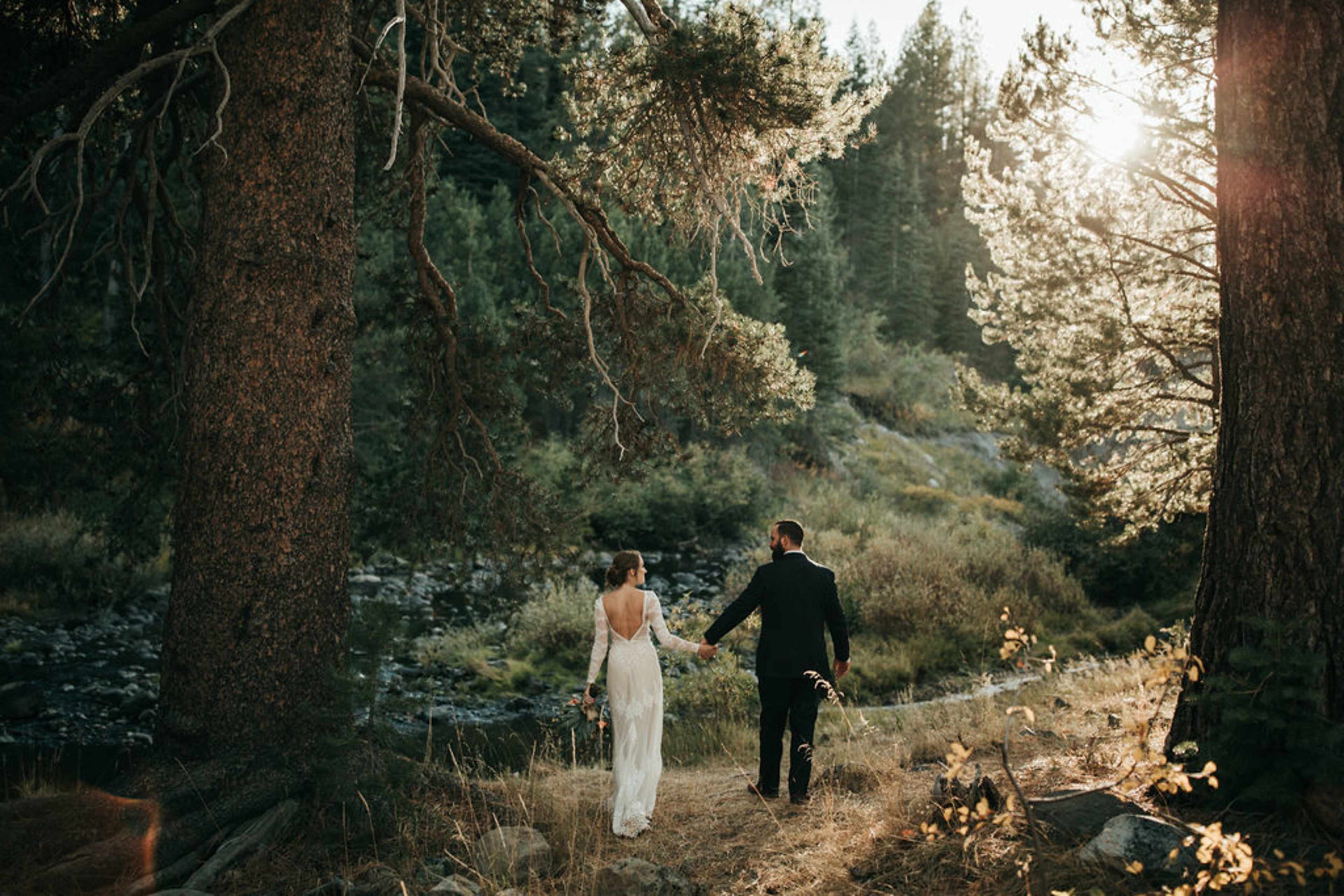 A bride and groom walk hand in hand along a forested path beside a stream, with sunlight filtering through the trees.