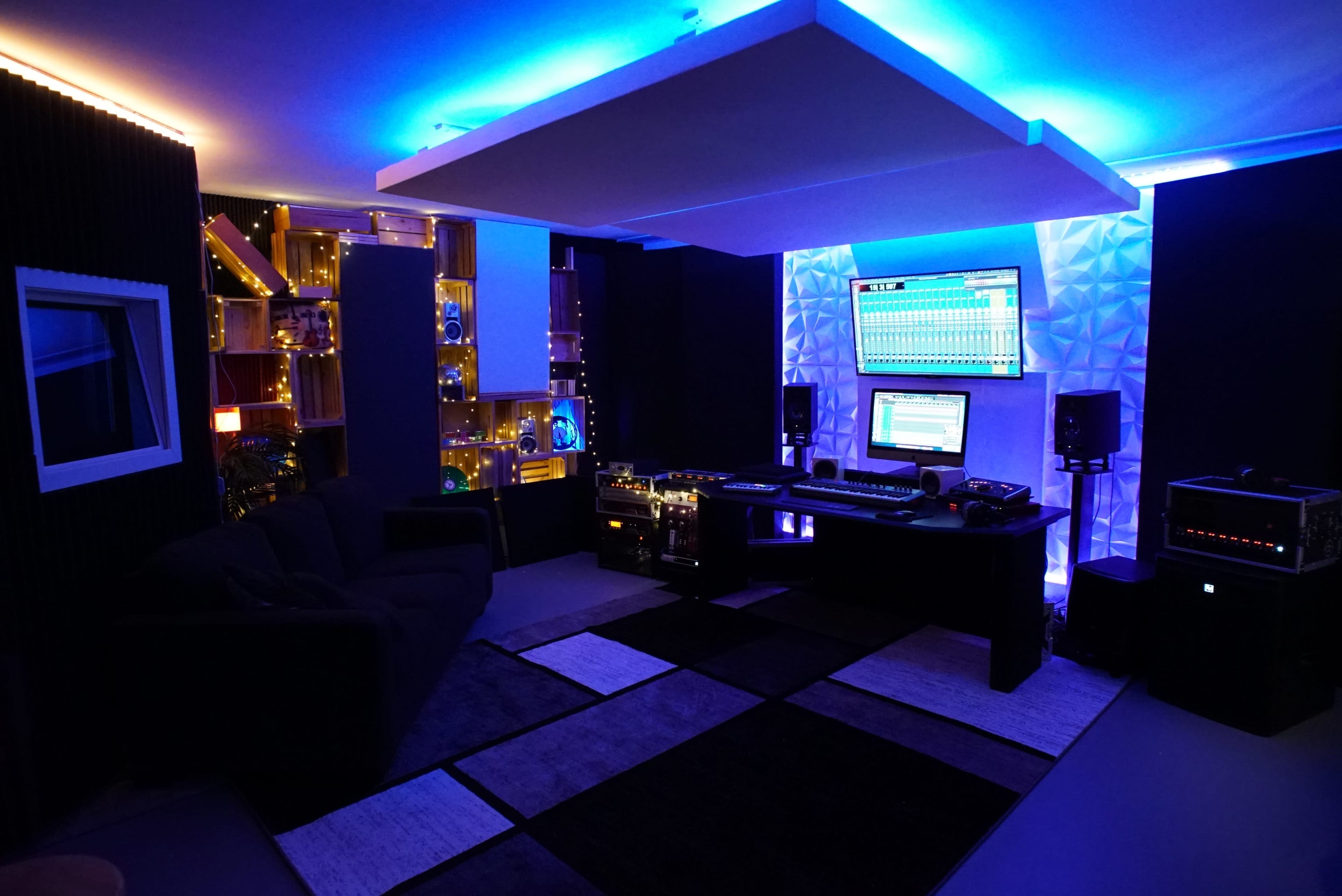 A modern music production studio features a black sofa, a large desk with audio equipment, and illuminated shelves, all bathed in blue and white lighting.