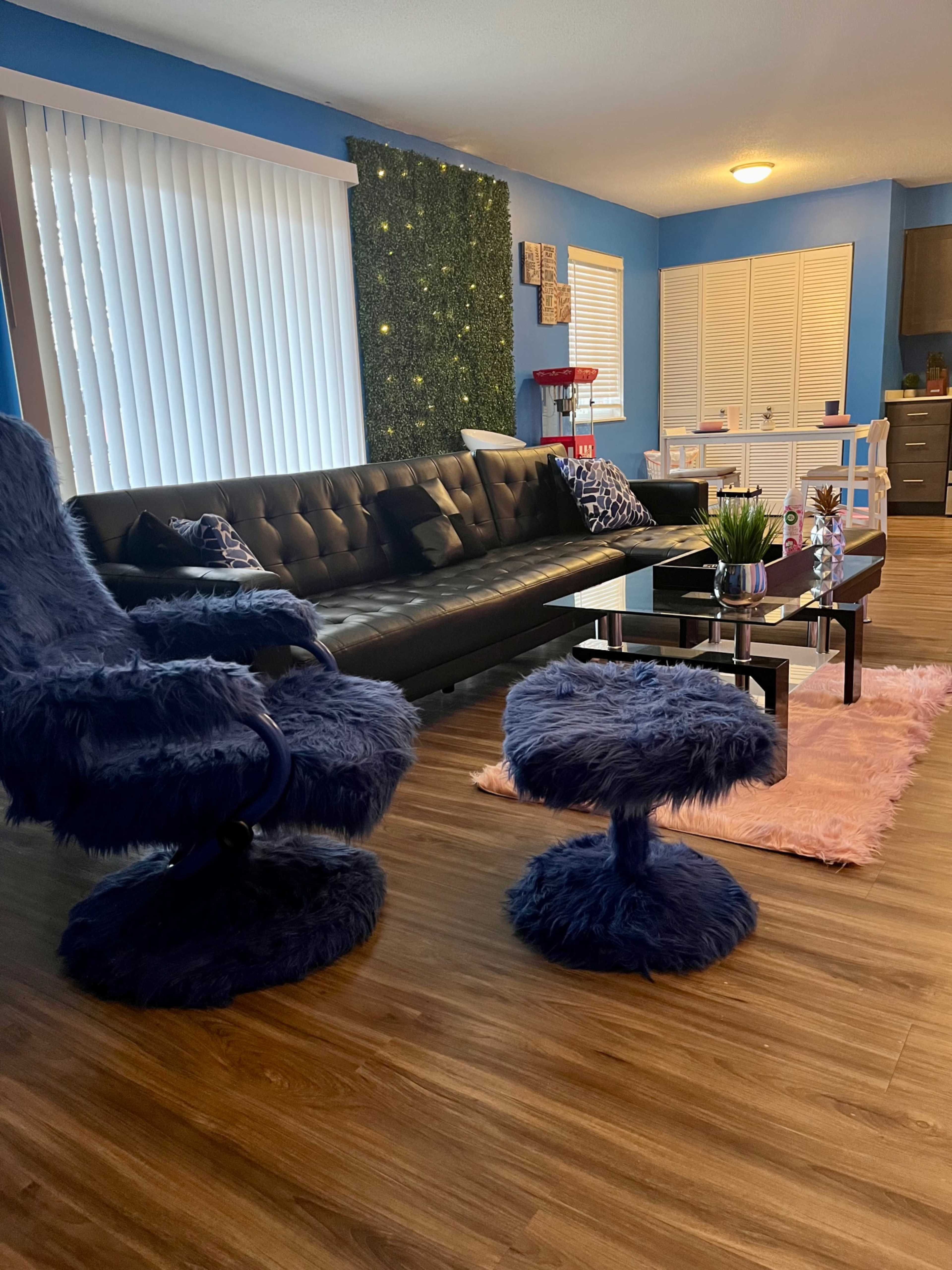 The living room features a black sofa, a blue, fuzzy armchair, and a matching ottoman, with wooden flooring and decorative elements on the walls.