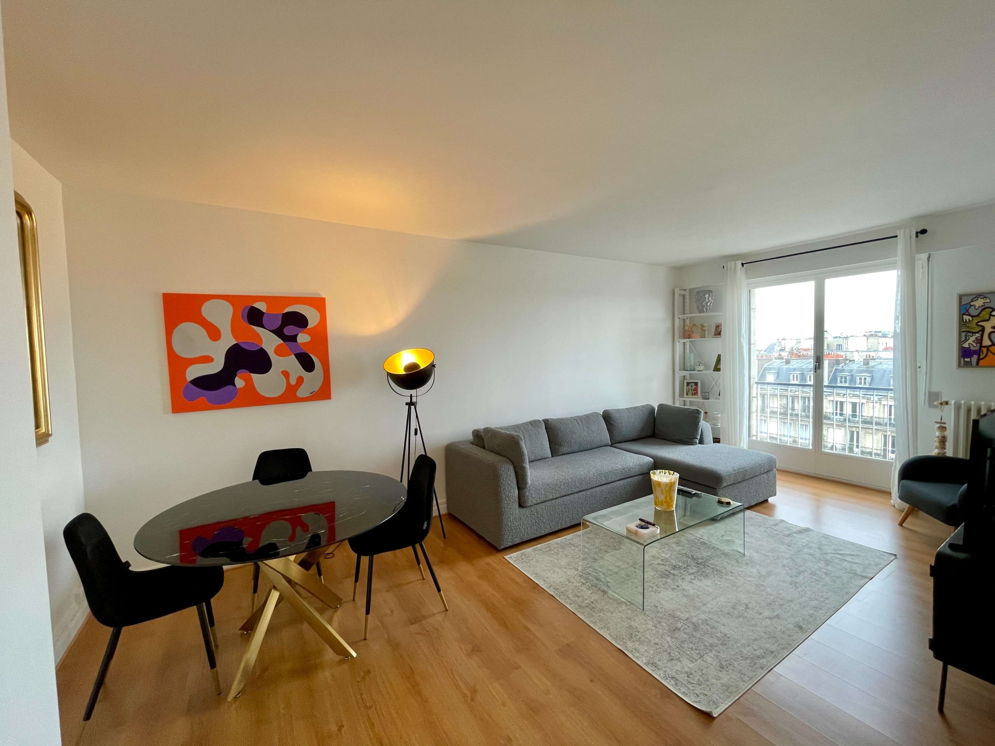 The image shows a modern living room with a gray couch, a round glass dining table, and a large window overlooking a cityscape.