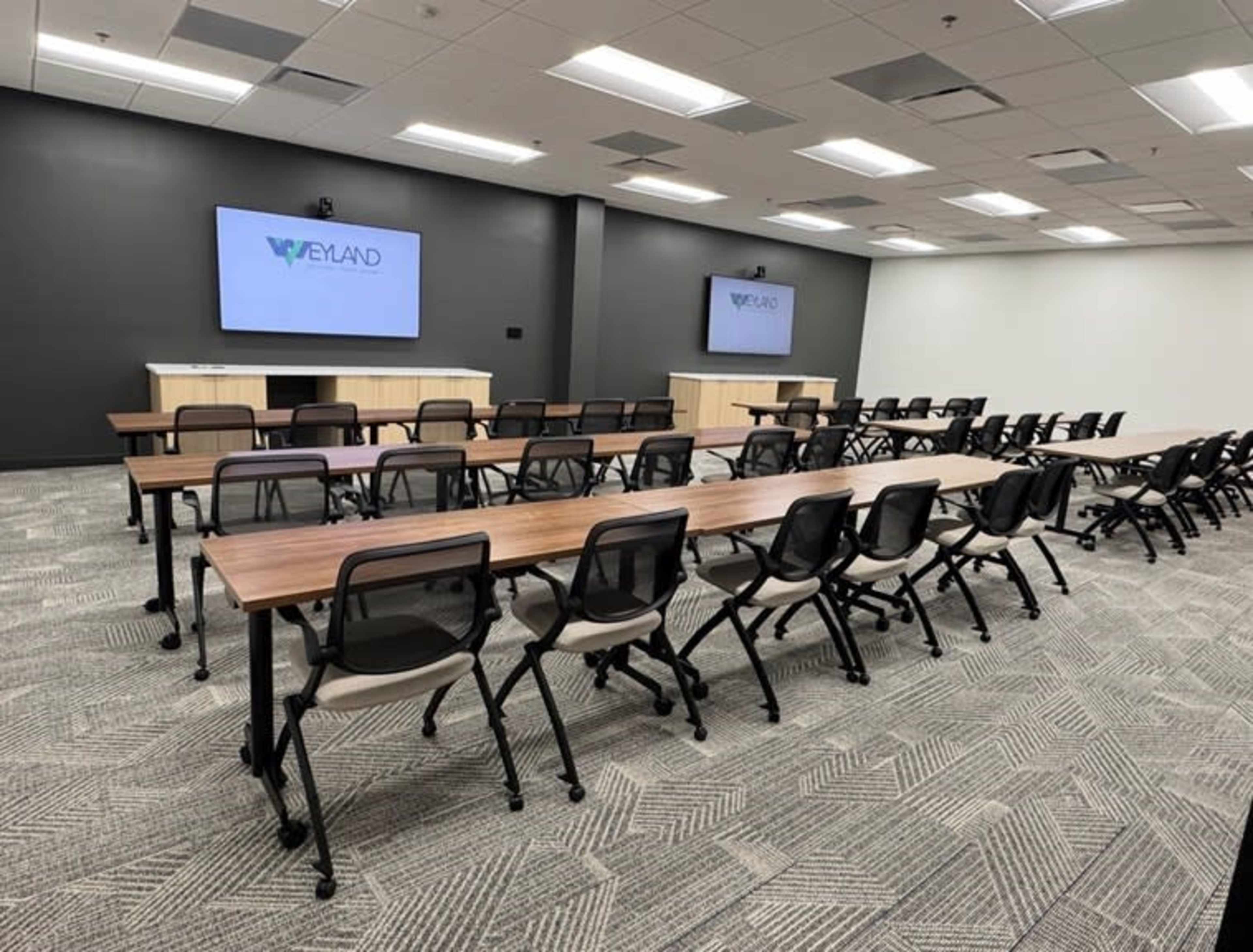 The image shows a modern conference room with rows of tables and chairs, two large screens on the front wall, and a dark-colored accent wall.