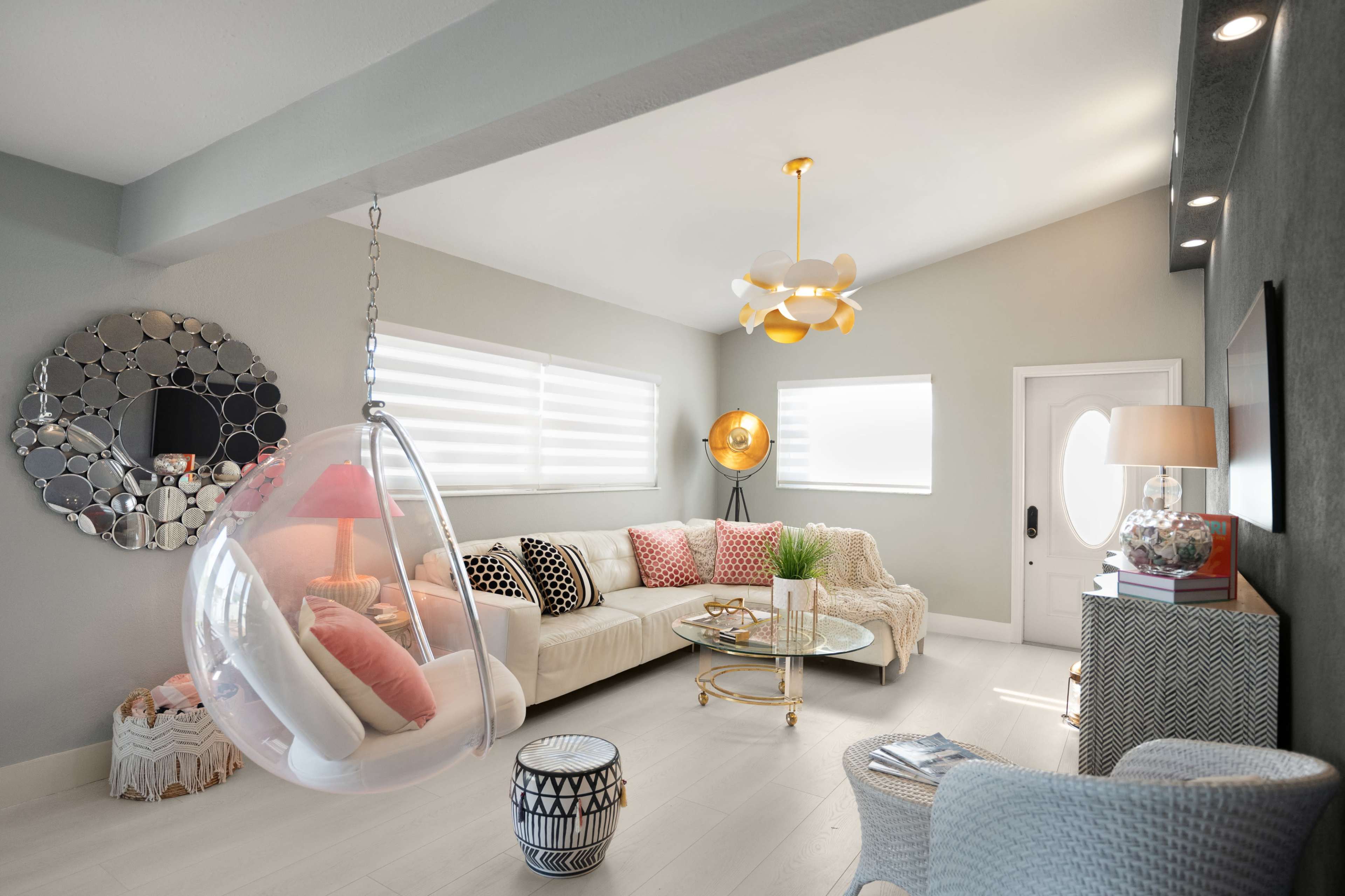 The living room features a modern transparent hanging chair, a light-colored sofa adorned with pink cushions, and a round glass coffee table, illuminated by a stylish ceiling light.