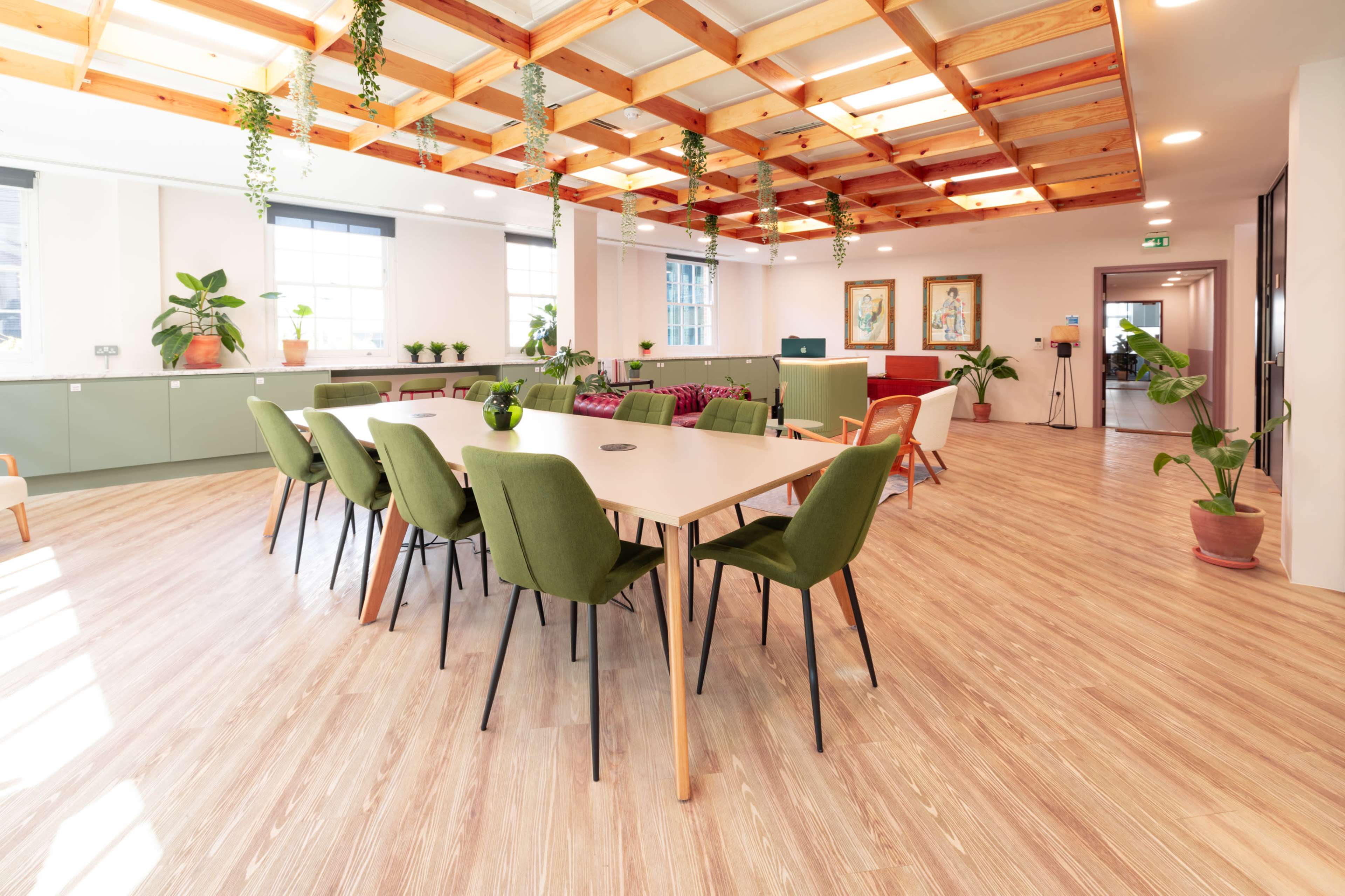 The image shows a modern office space with a long rectangular table surrounded by green chairs, large windows, and plants integrated into the decor.