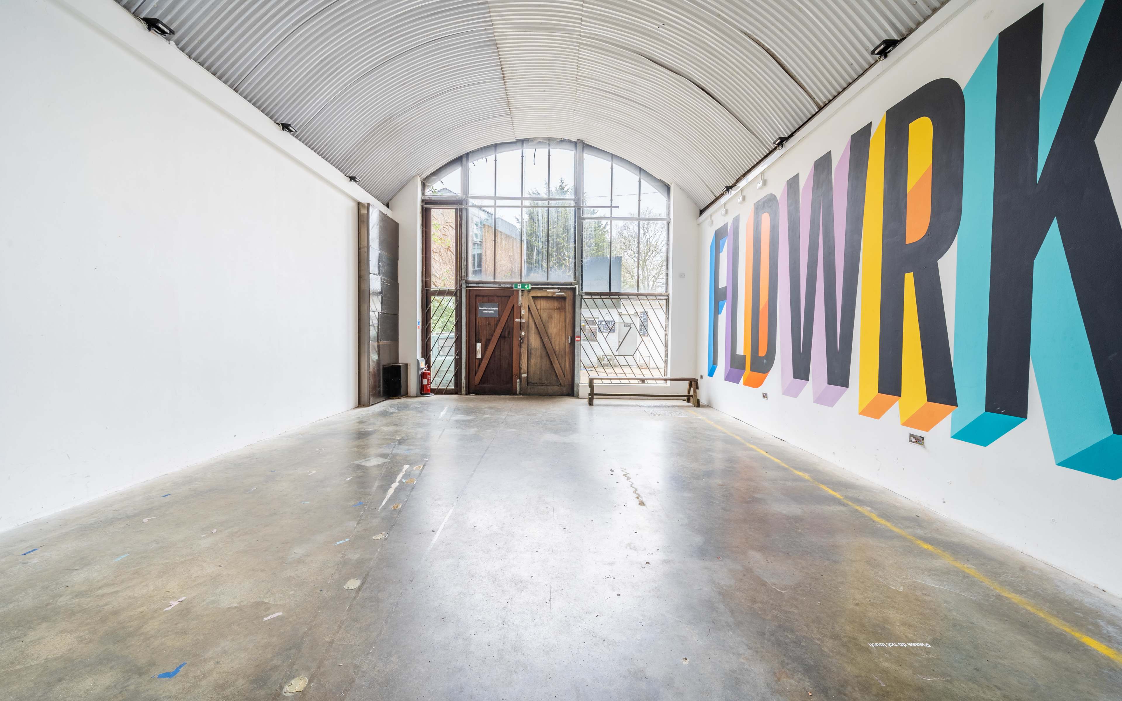 A spacious, minimalist interior with a large colorful mural that reads "FIDWRK" on one wall and a prominent glass door at the entrance.