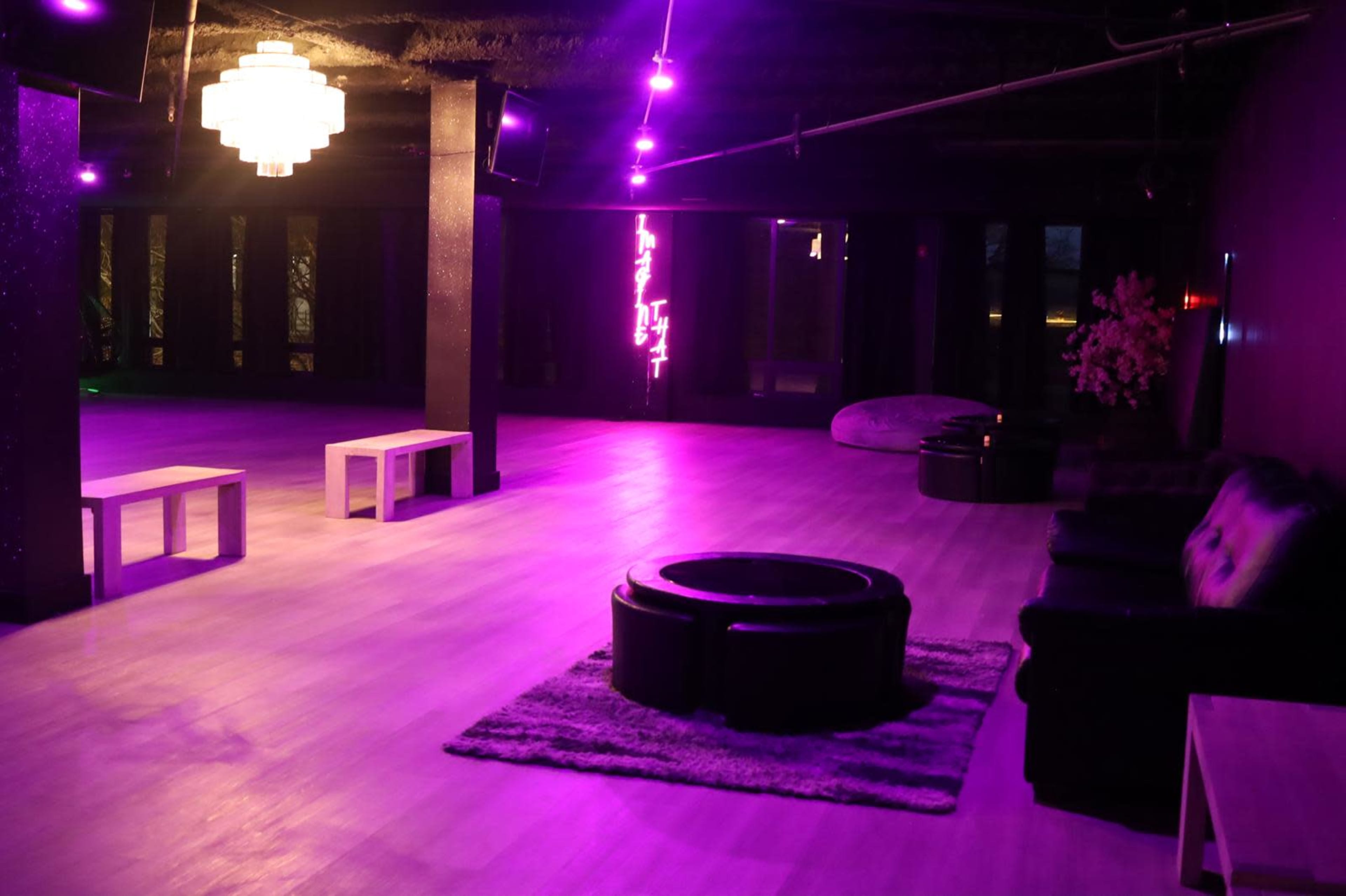 The image shows a dimly lit lounge area with purple lighting, featuring seating, low tables, and decorative elements in a spacious, empty room.