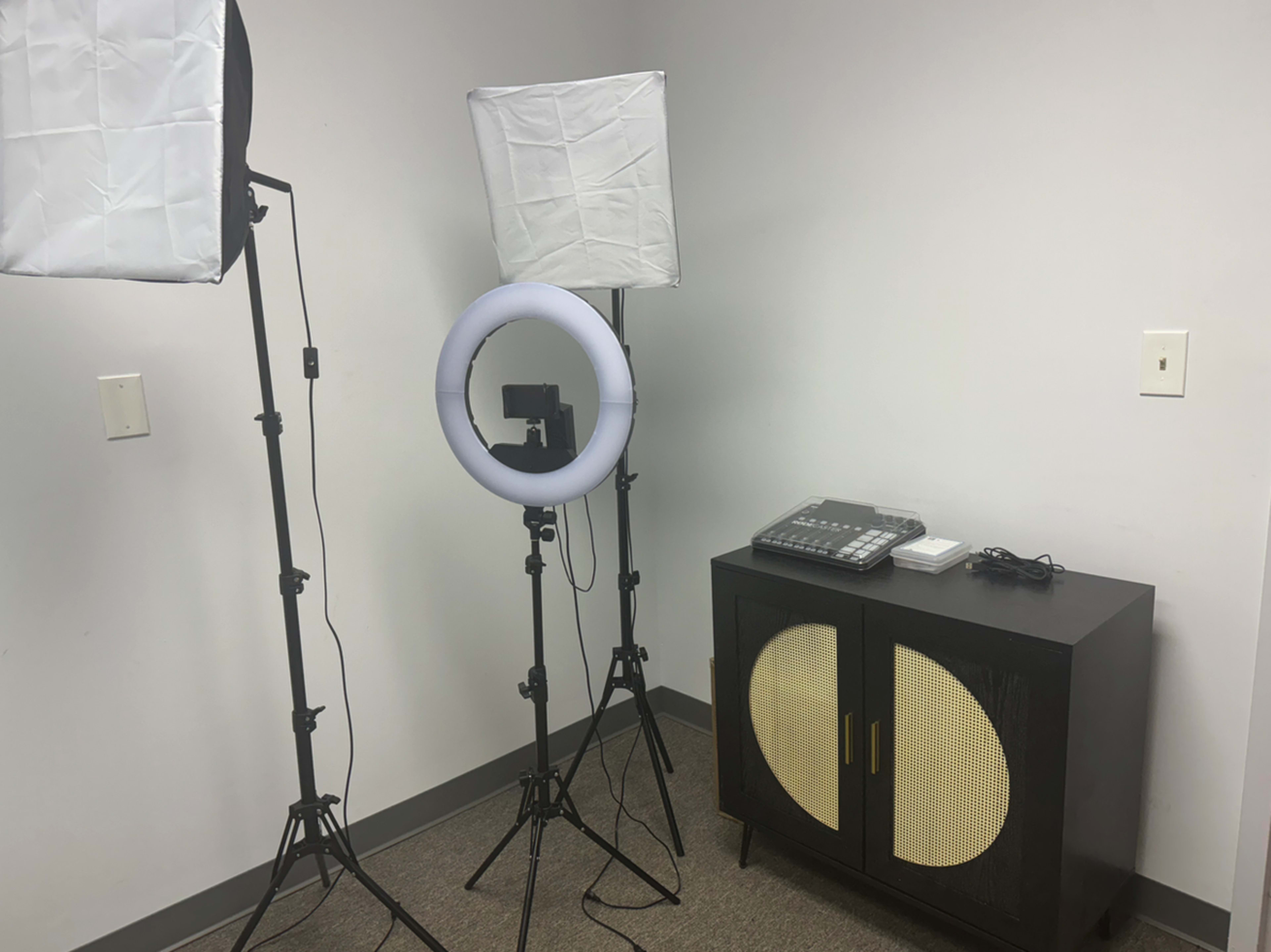 The scene shows a minimalistic setup with two softbox lights and a ring light positioned near a black cabinet and a phone on a carpeted floor in a room with white walls.