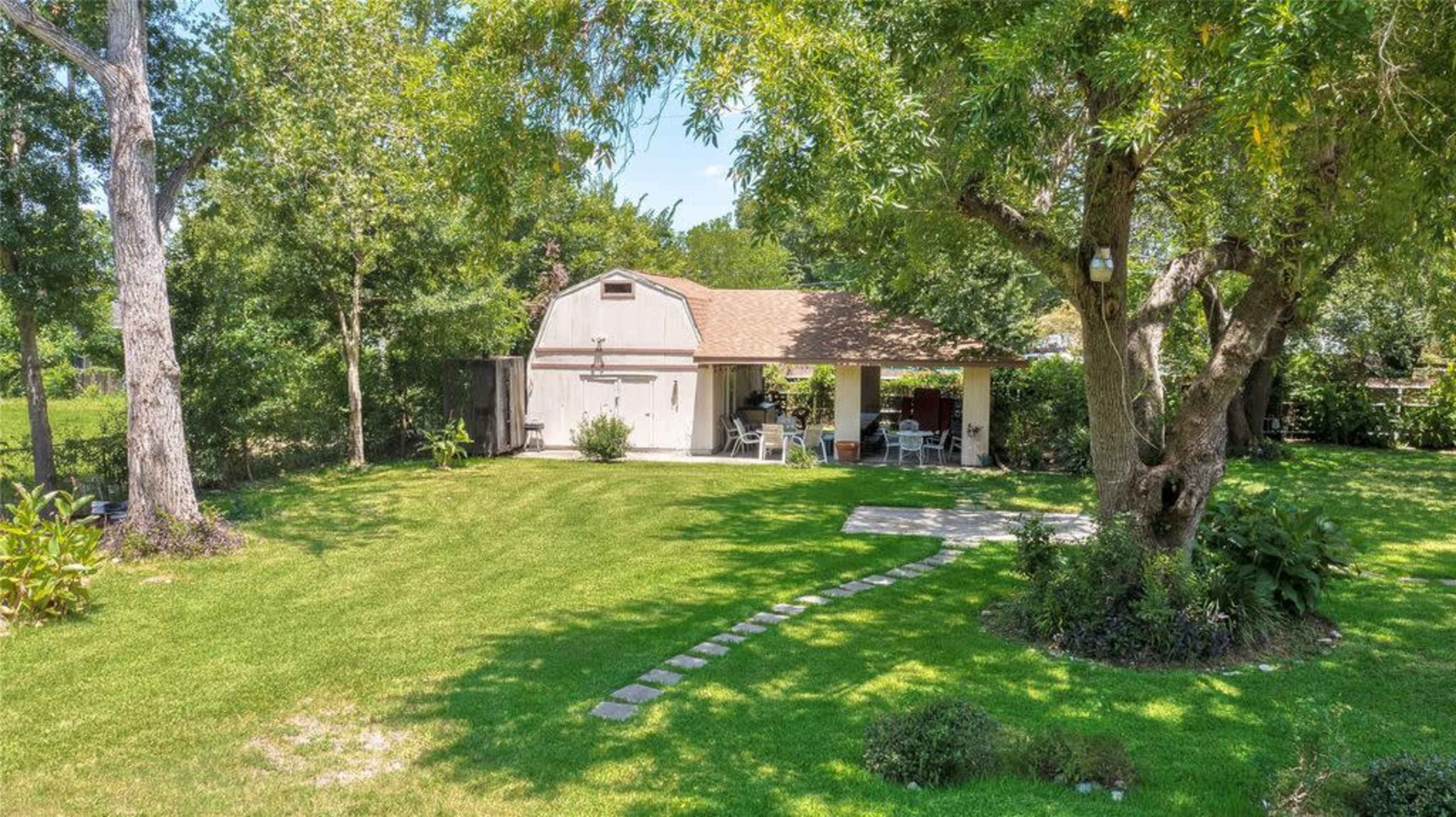 A small, single-story house with a brown roof is situated among trees, featuring a stone pathway leading to a patio area.