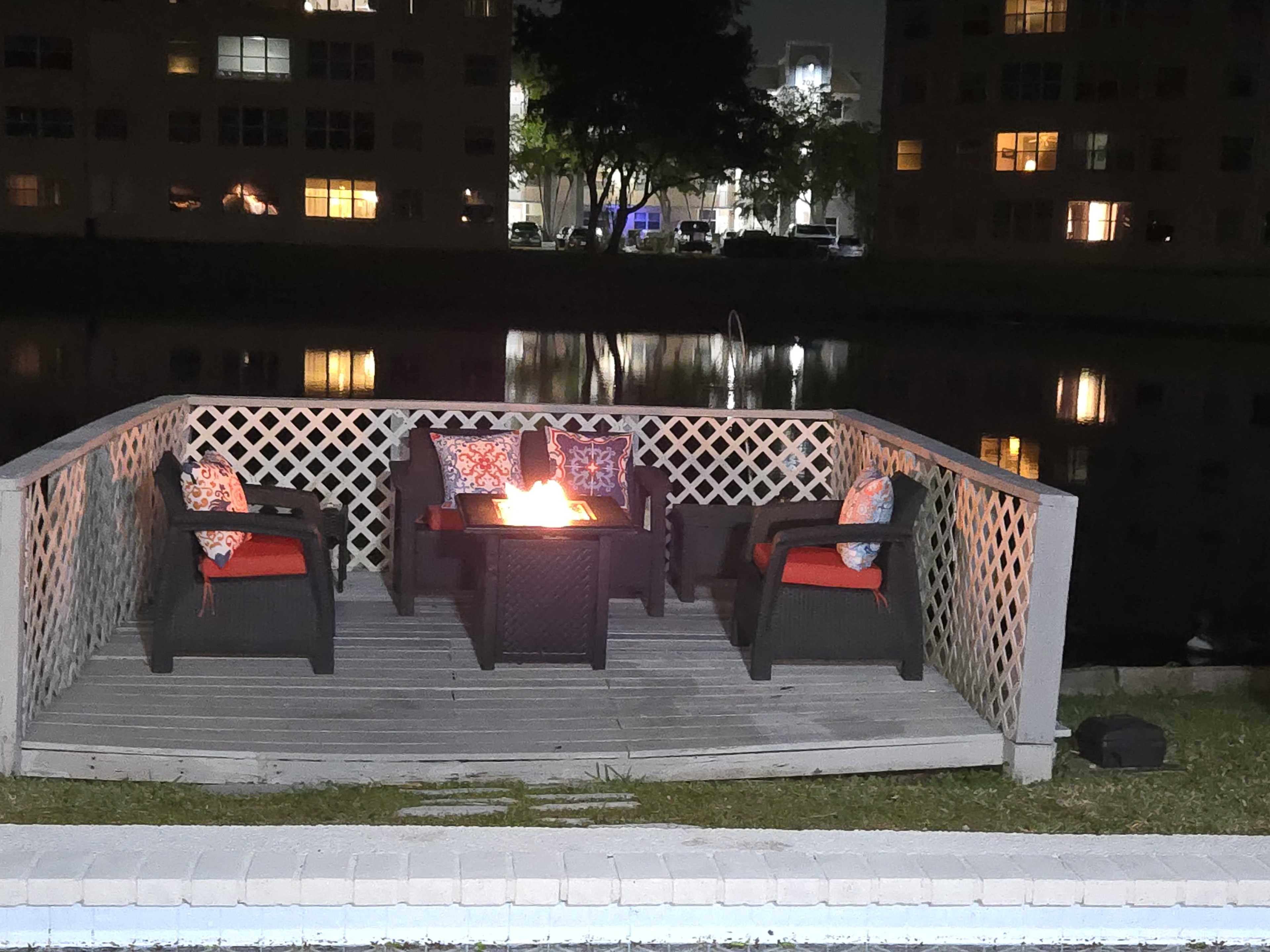 A small wooden deck by a pond features four chairs with cushions arranged around a fire pit, illuminated by the fire at night.