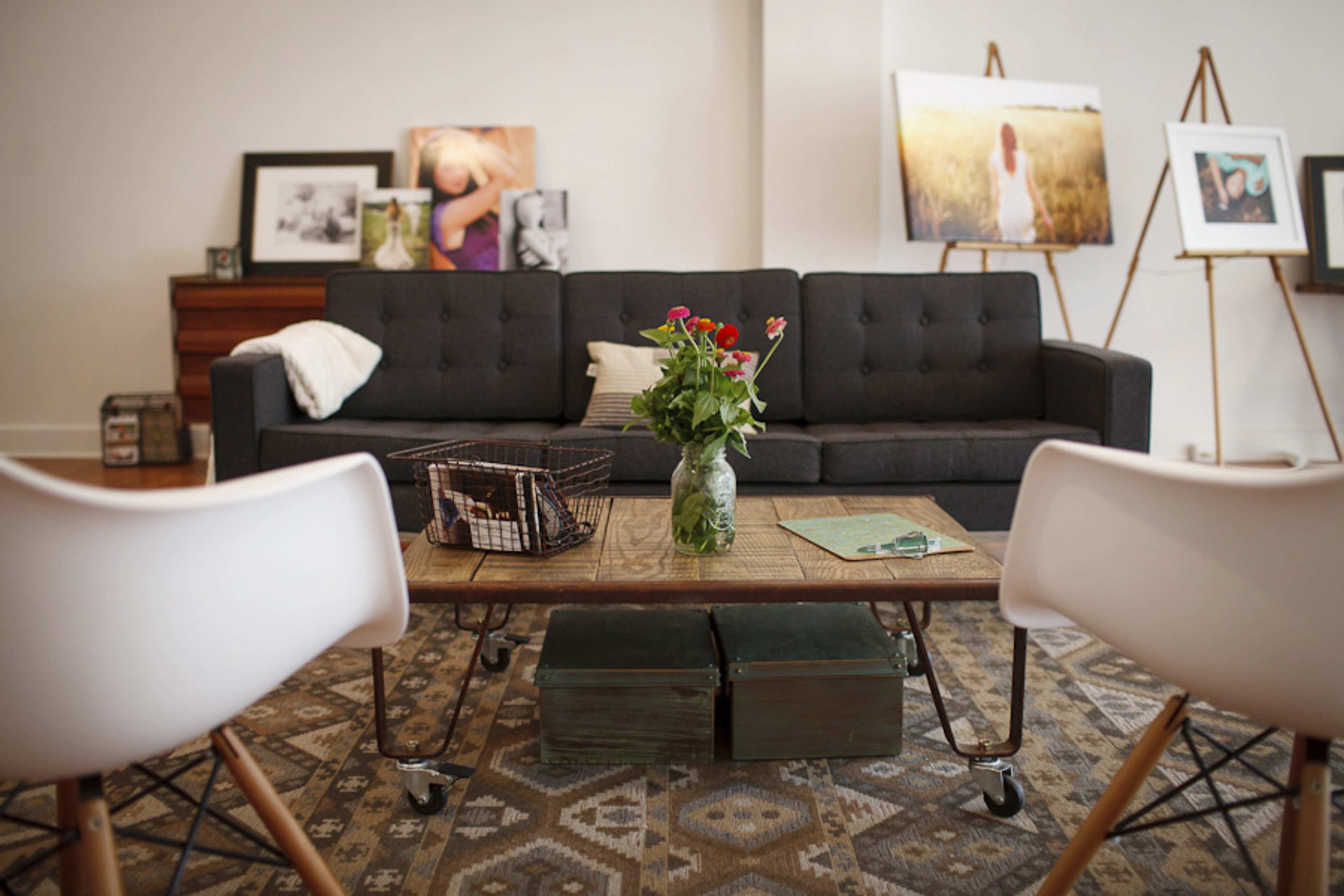 A modern living room with a gray sofa, a wooden coffee table with a vase of flowers, and two white chairs, surrounded by framed photos and artwork on easels.