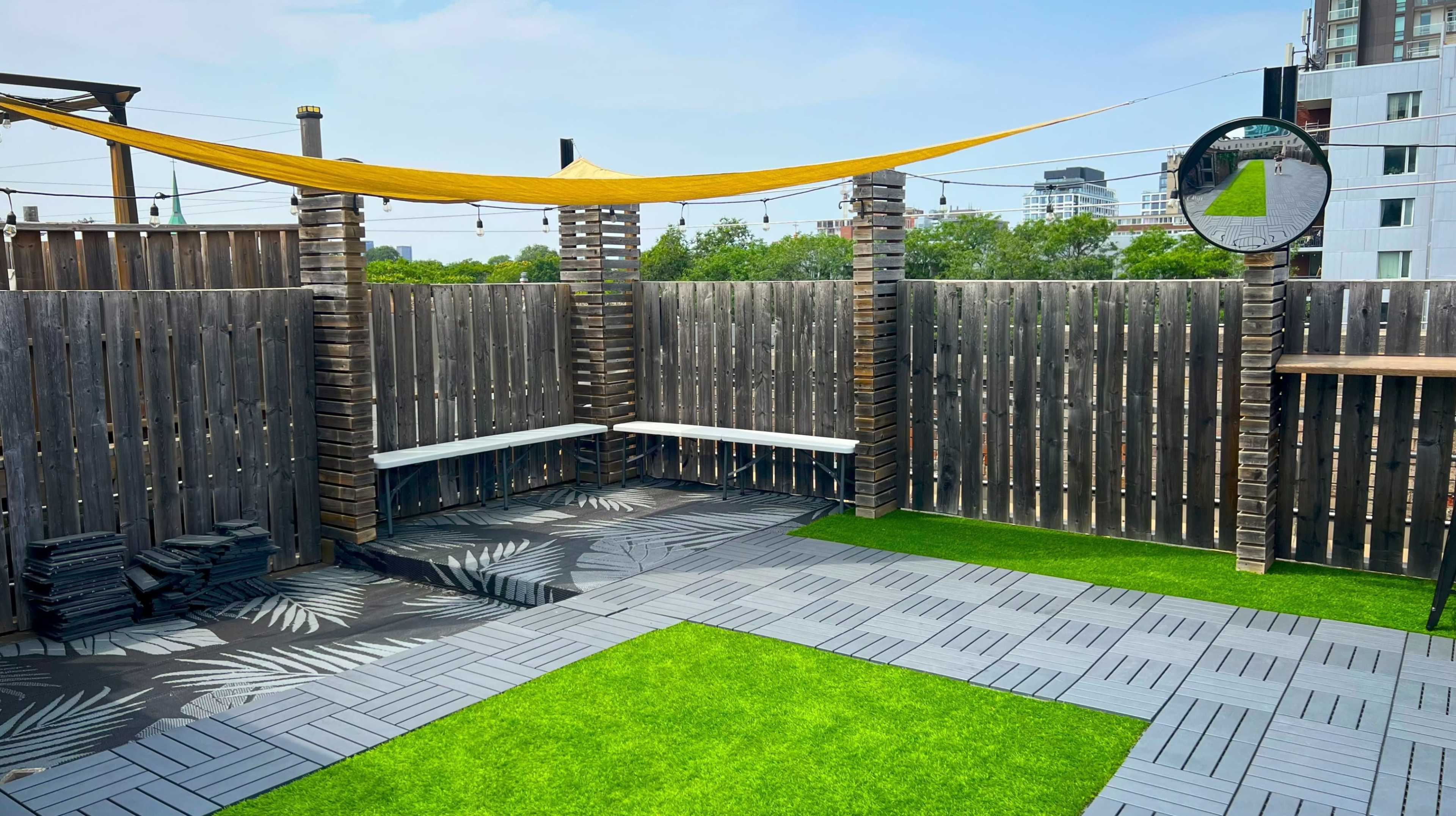 The image shows a rooftop space with a grass-like surface, wooden fencing, and a yellow fabric shade stretched between two pillars.