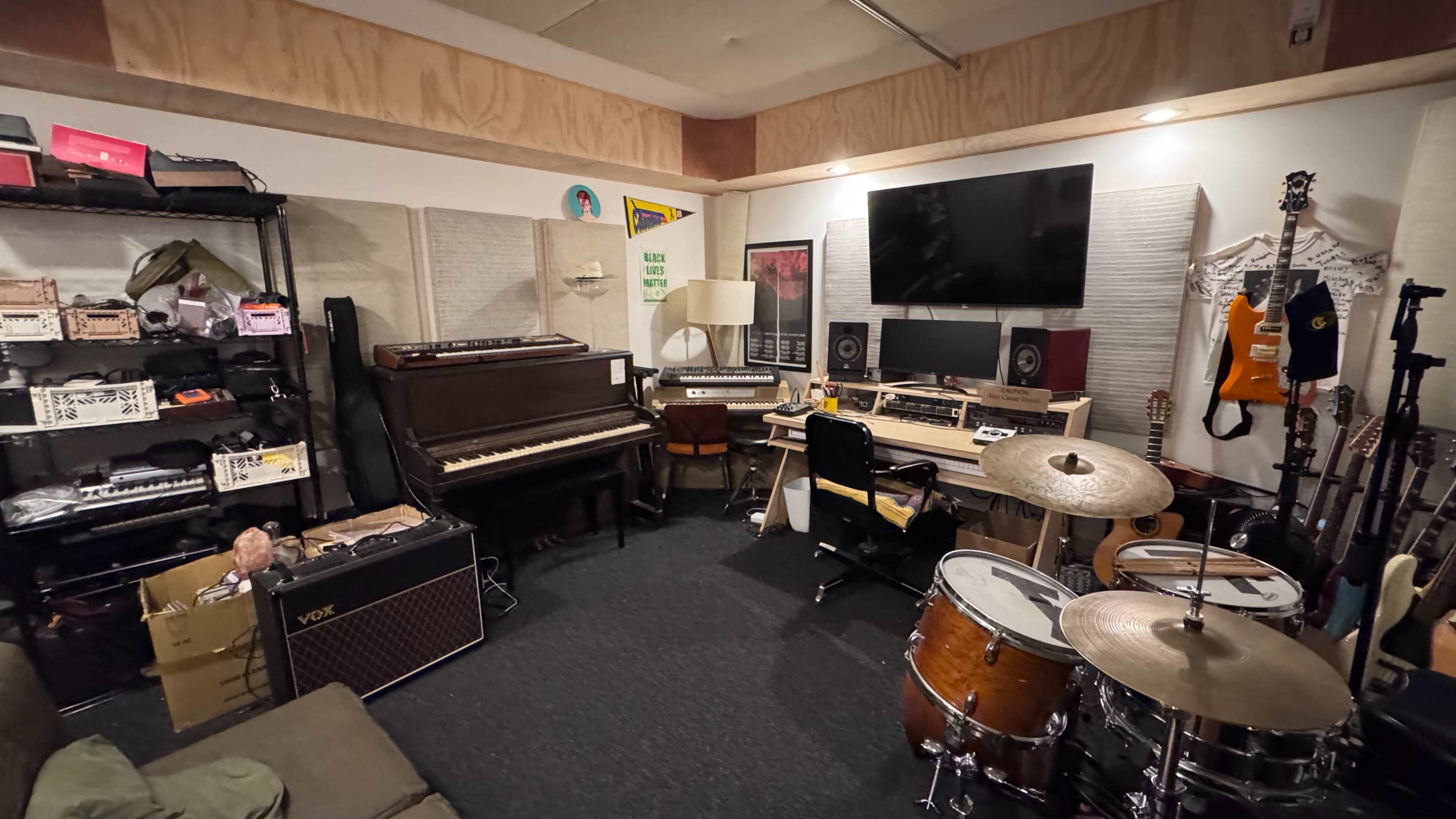 The image shows a small music studio featuring various instruments, including a drum set, a piano, and several guitars, along with sound equipment and storage shelves.