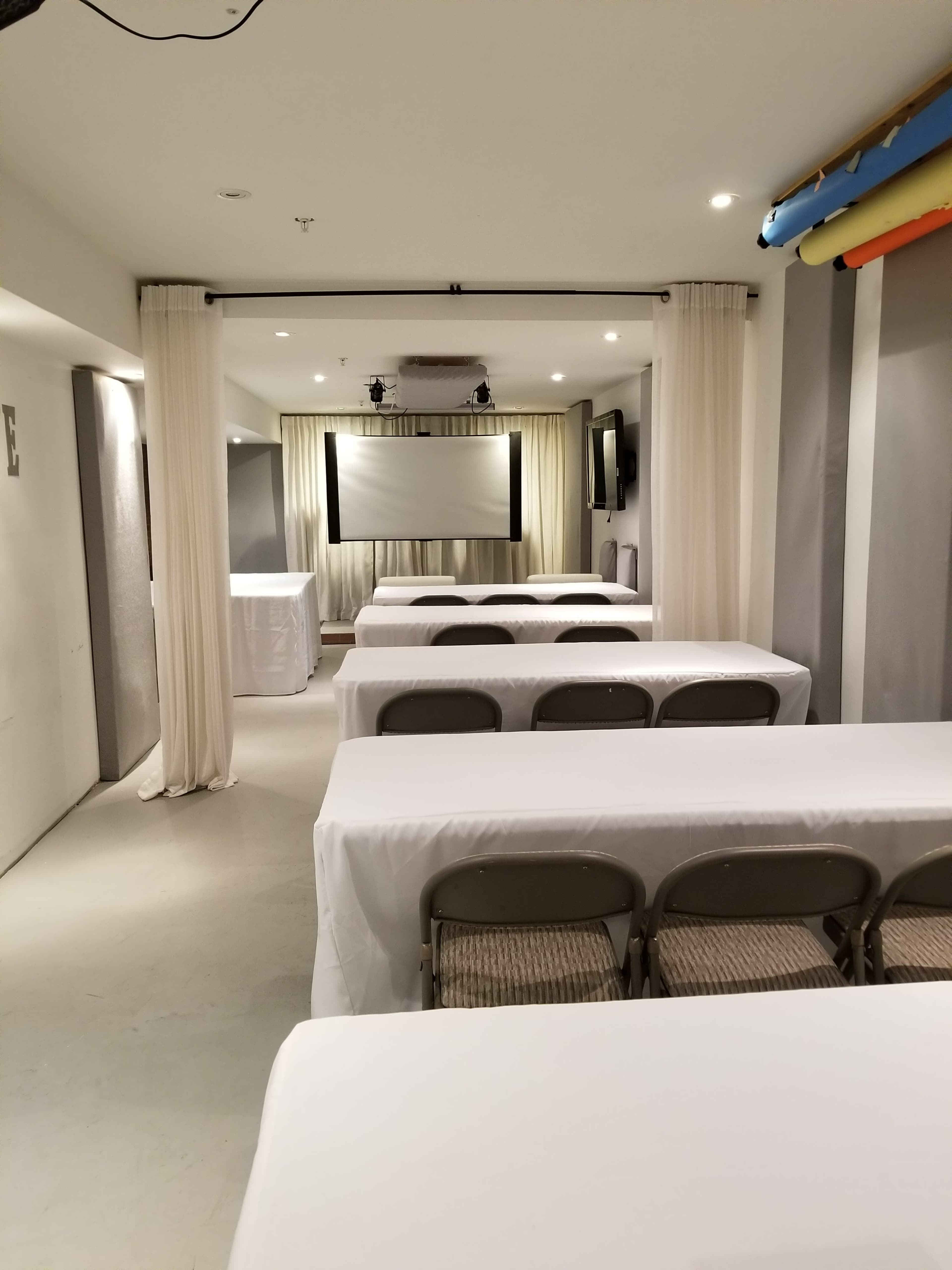 A conference room features several rows of tables covered with white tablecloths, facing a projector screen at the front.
