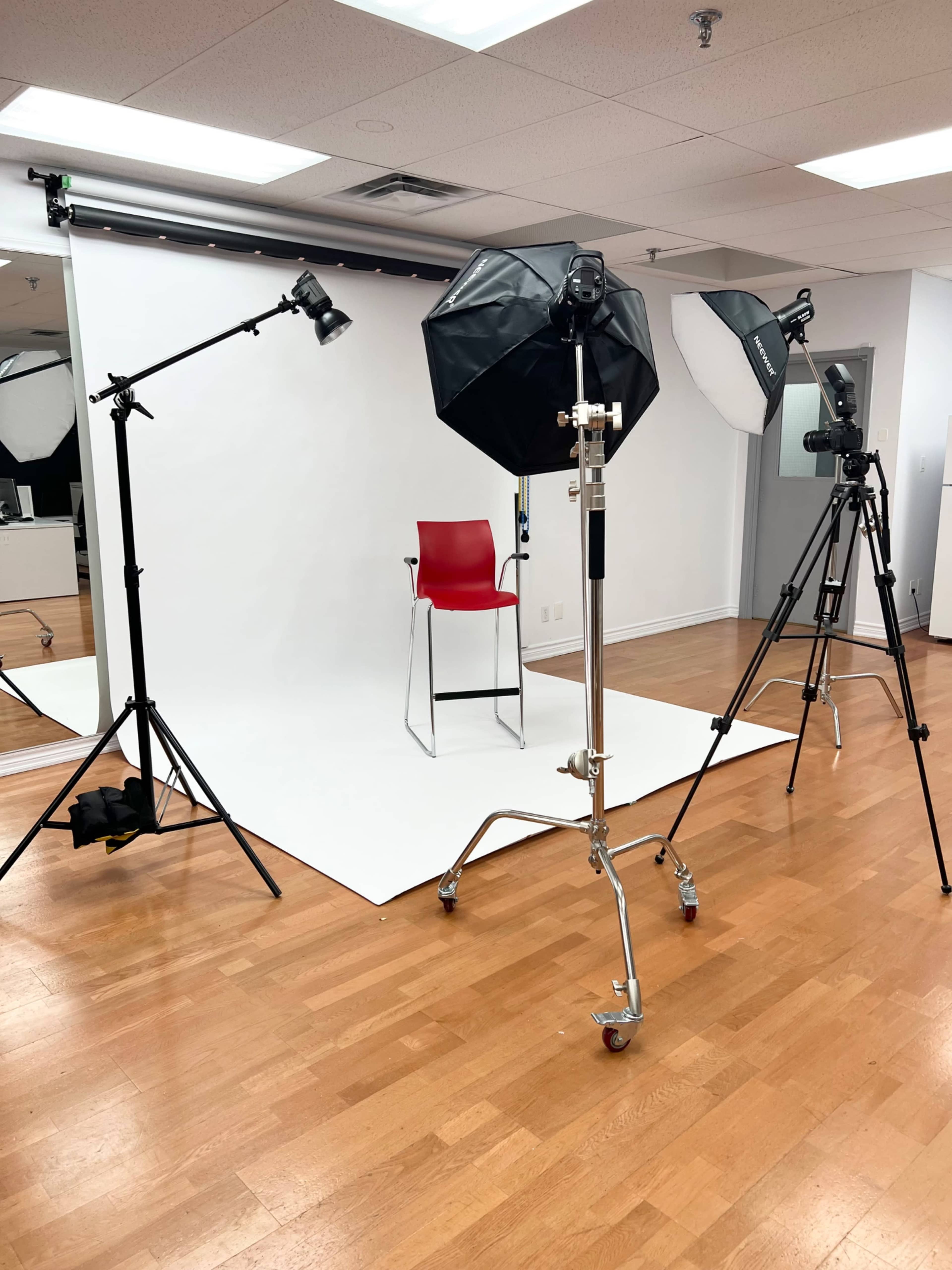 The image shows a photography studio setup with lighting equipment, a camera on a tripod, and a red chair on a white backdrop.