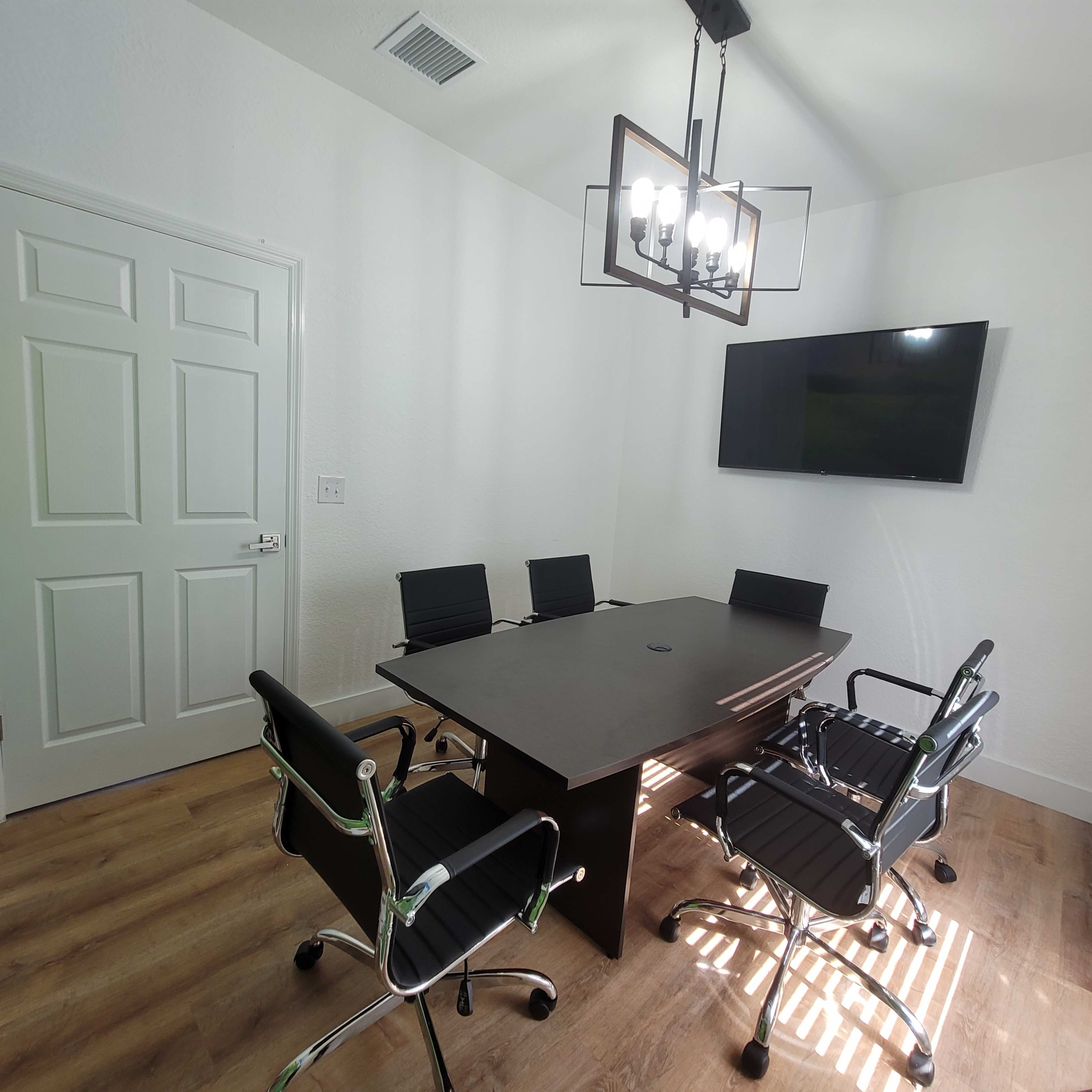 A conference room featuring a rectangular black table surrounded by four black chairs, a television on the wall, and a ceiling light fixture.