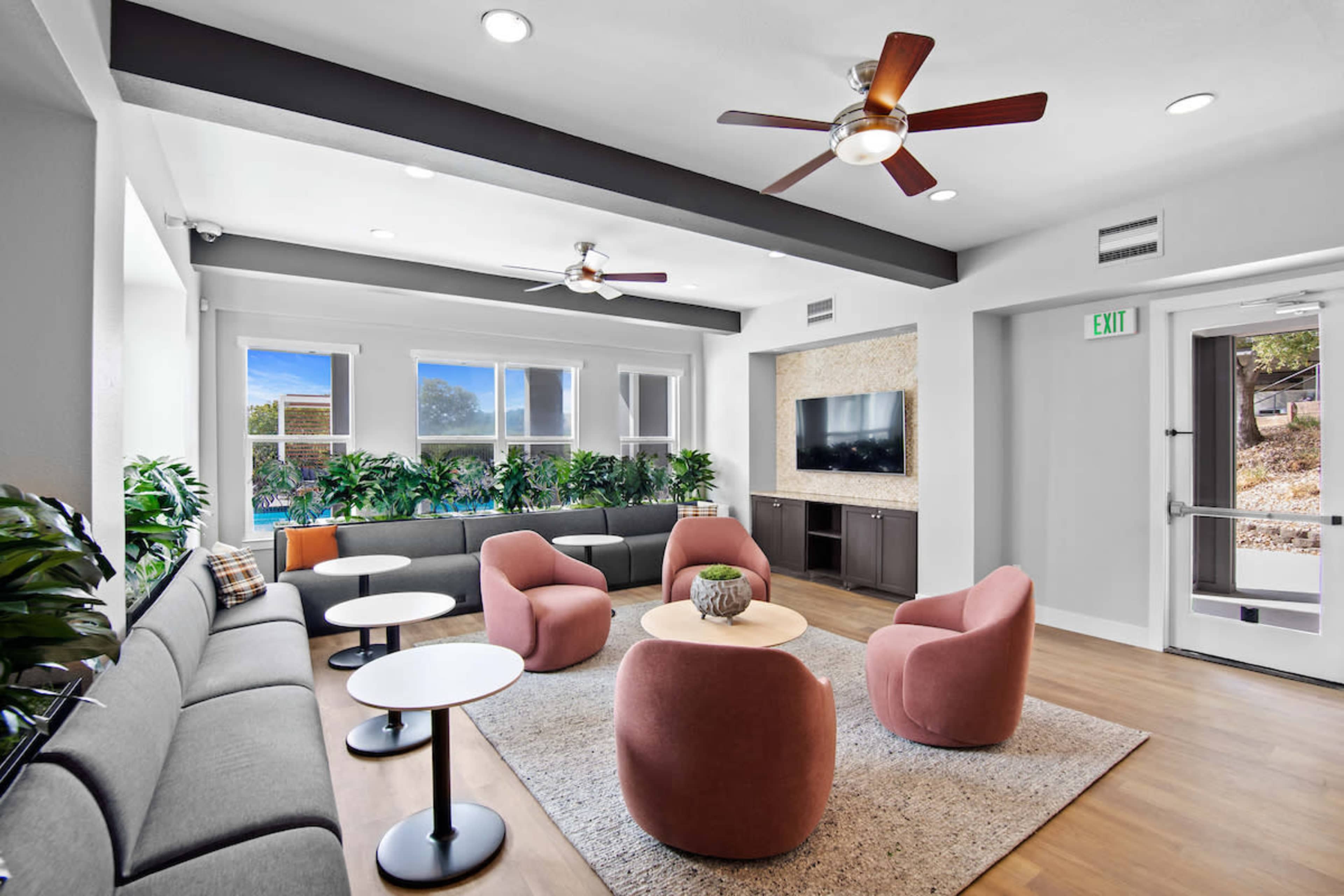 The image shows a modern lounge area featuring a sectional sofa, several round coffee tables, and pink upholstered chairs, with large windows and indoor plants enhancing the space.