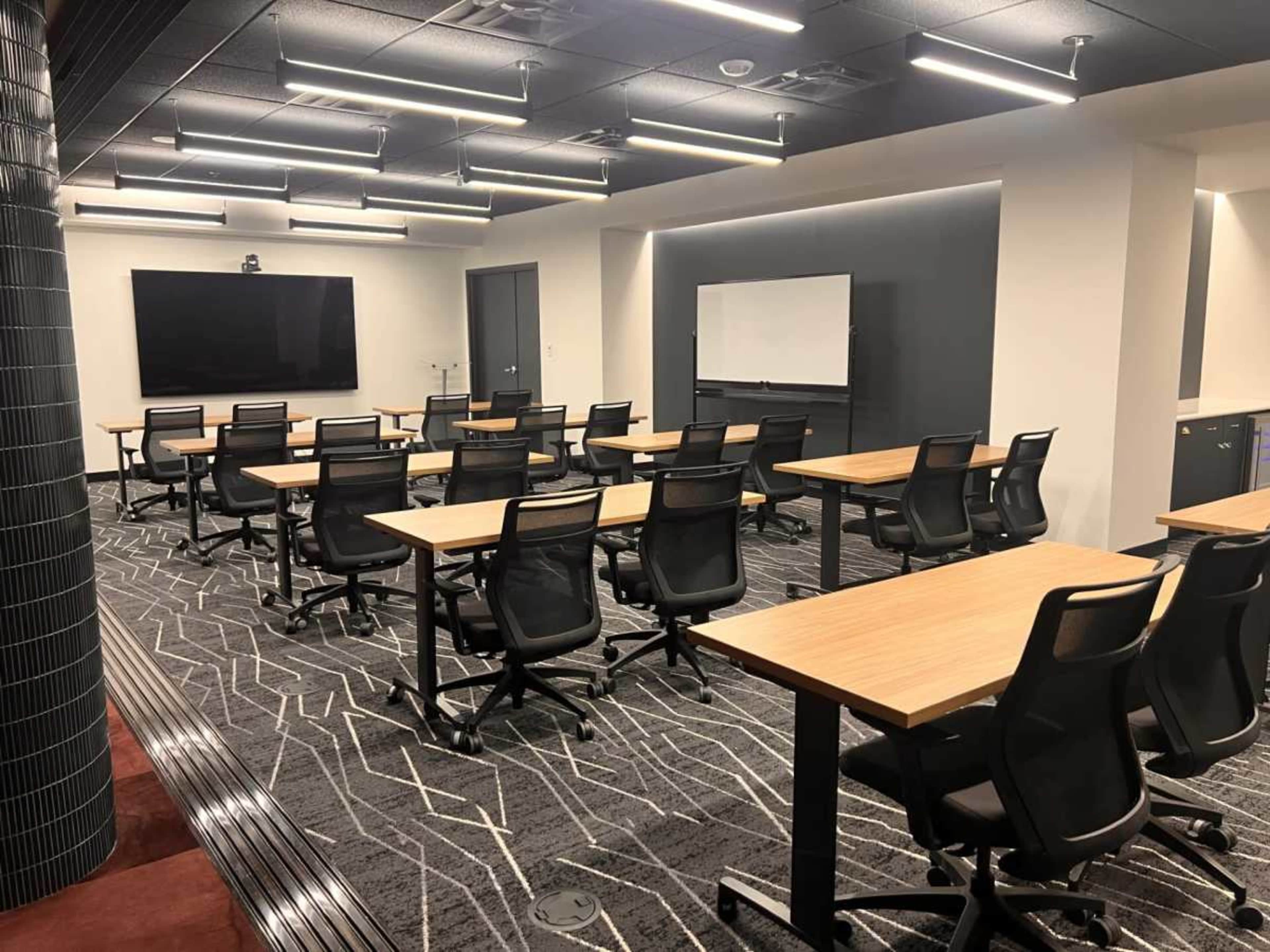 The image shows a modern conference room with rows of wooden tables and black ergonomic chairs arranged facing a large screen and a whiteboard.