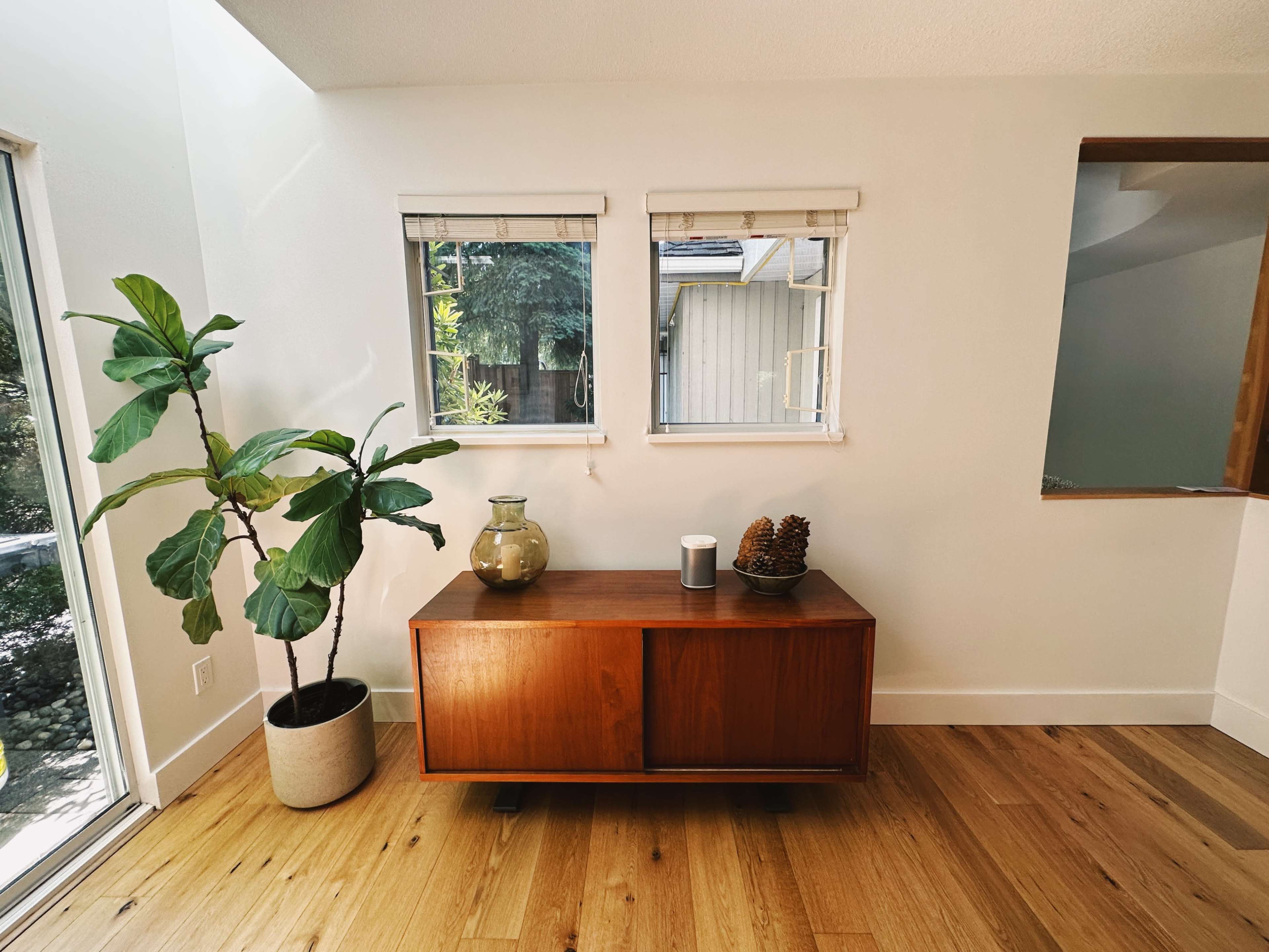 A wooden sideboard with a plant and decor items sits beneath two windows in a well-lit room.