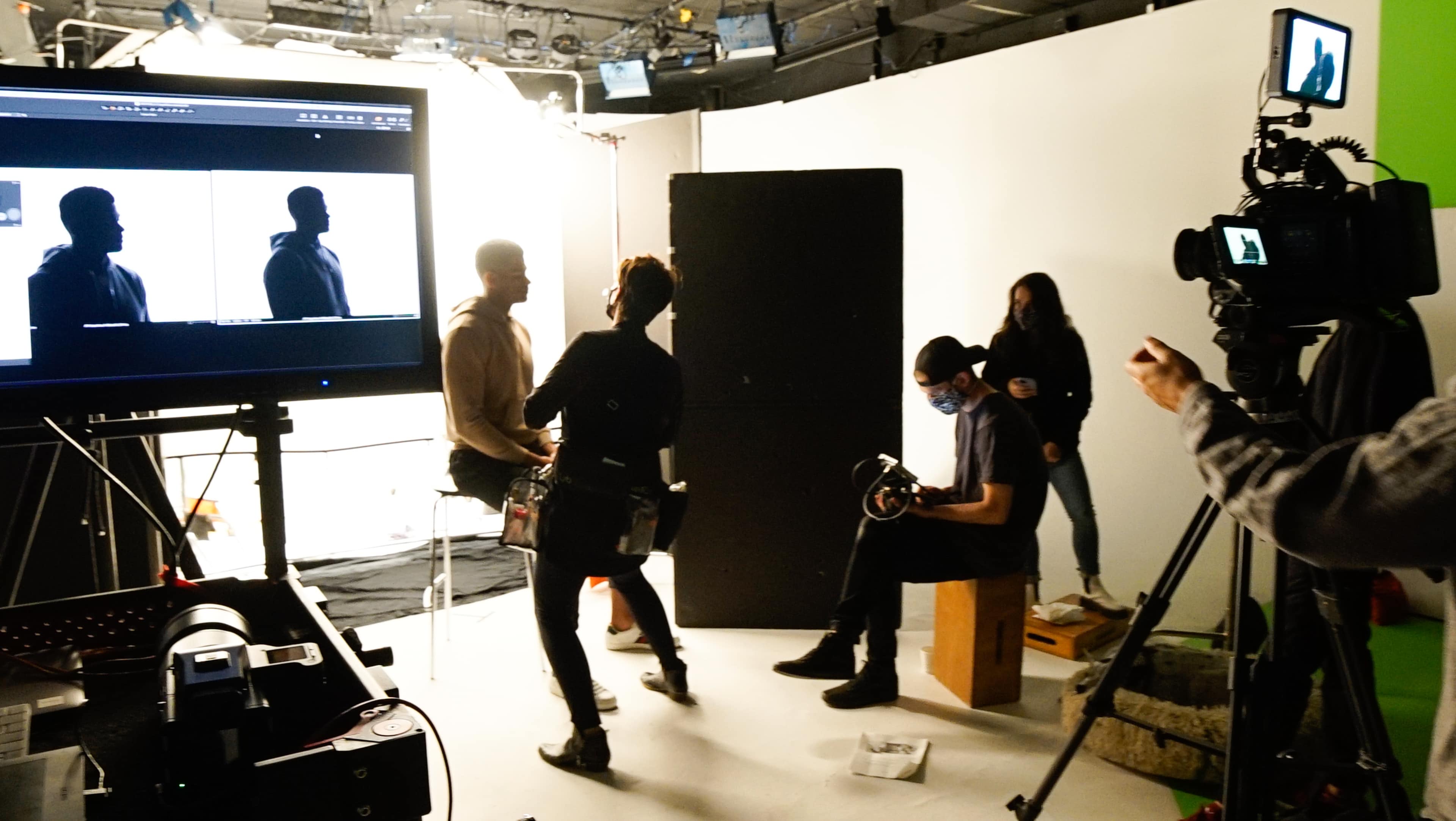 A film crew is preparing for a shoot in a studio, with several people working around a large monitor displaying silhouettes.
