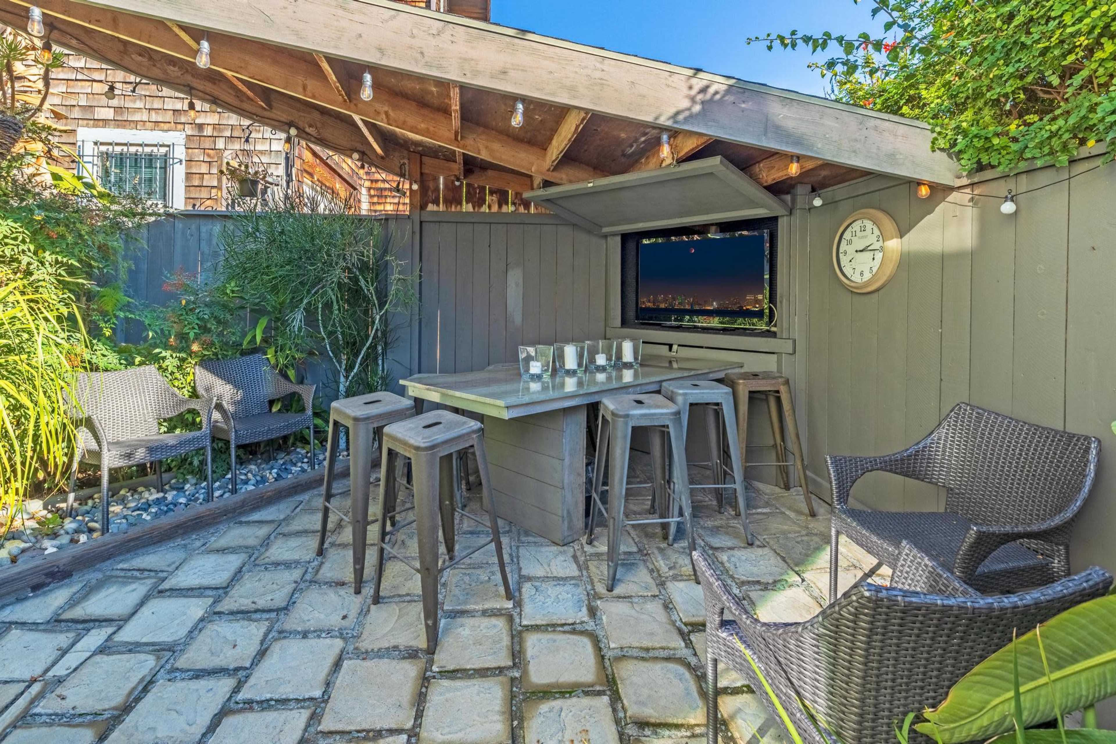 The image shows an outdoor bar area with a stone patio, surrounded by greenery and featuring high stools and chairs under a wooden roof.