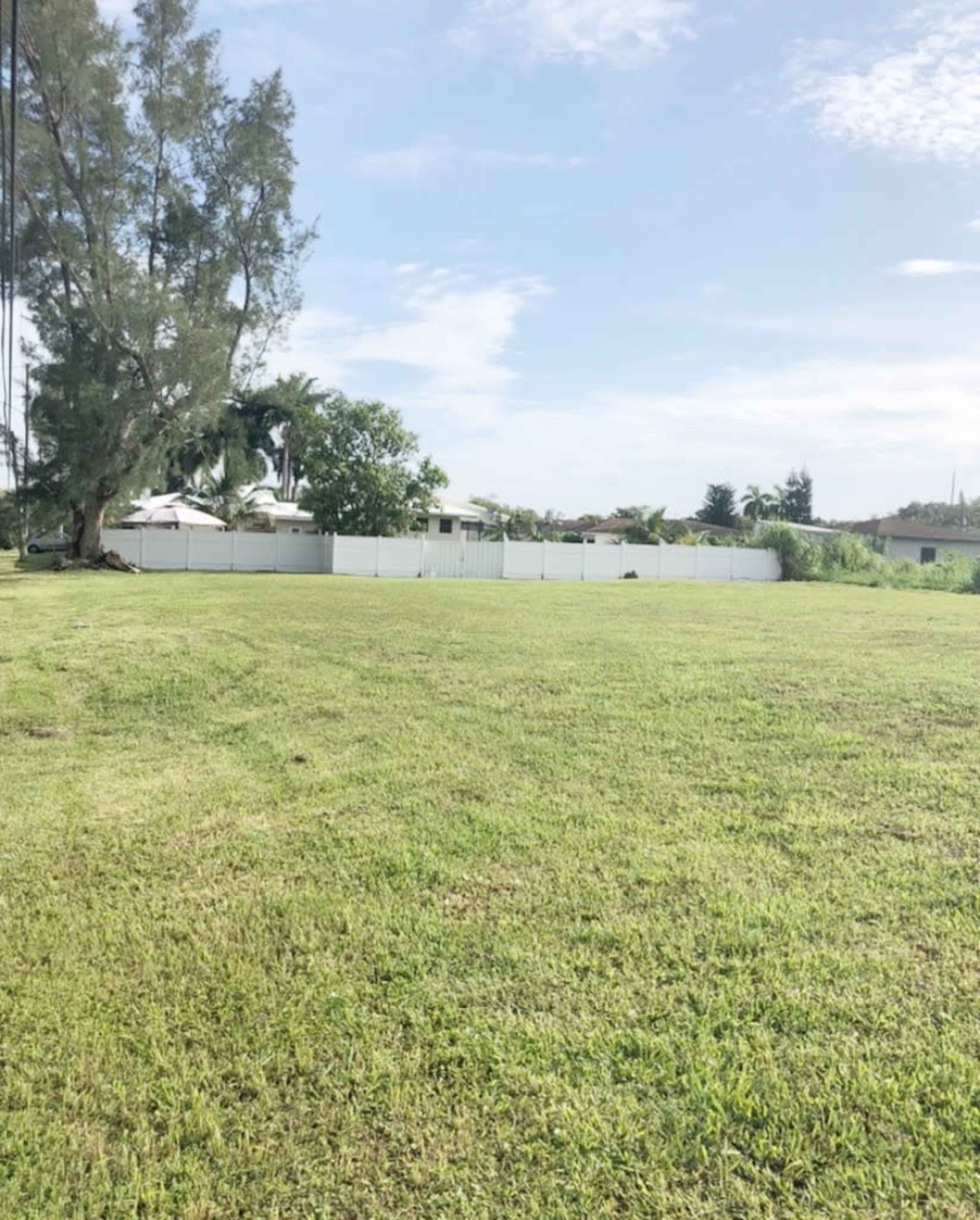 A large, grassy field bordered by a white fence with a few houses and trees in the background.