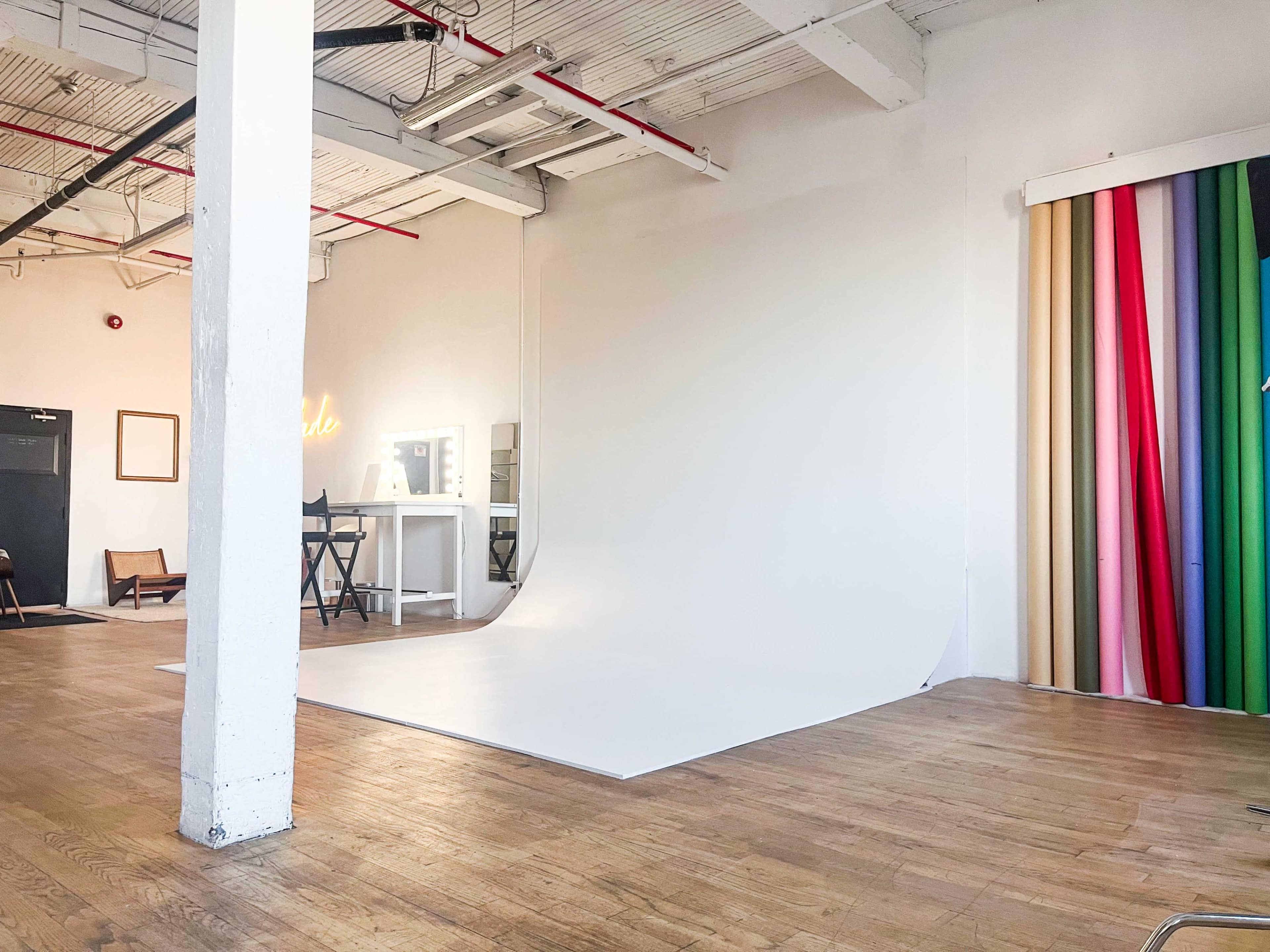 A spacious studio with a curved white backdrop, a mirror and table, and a colorful curtain arrangement on one side.