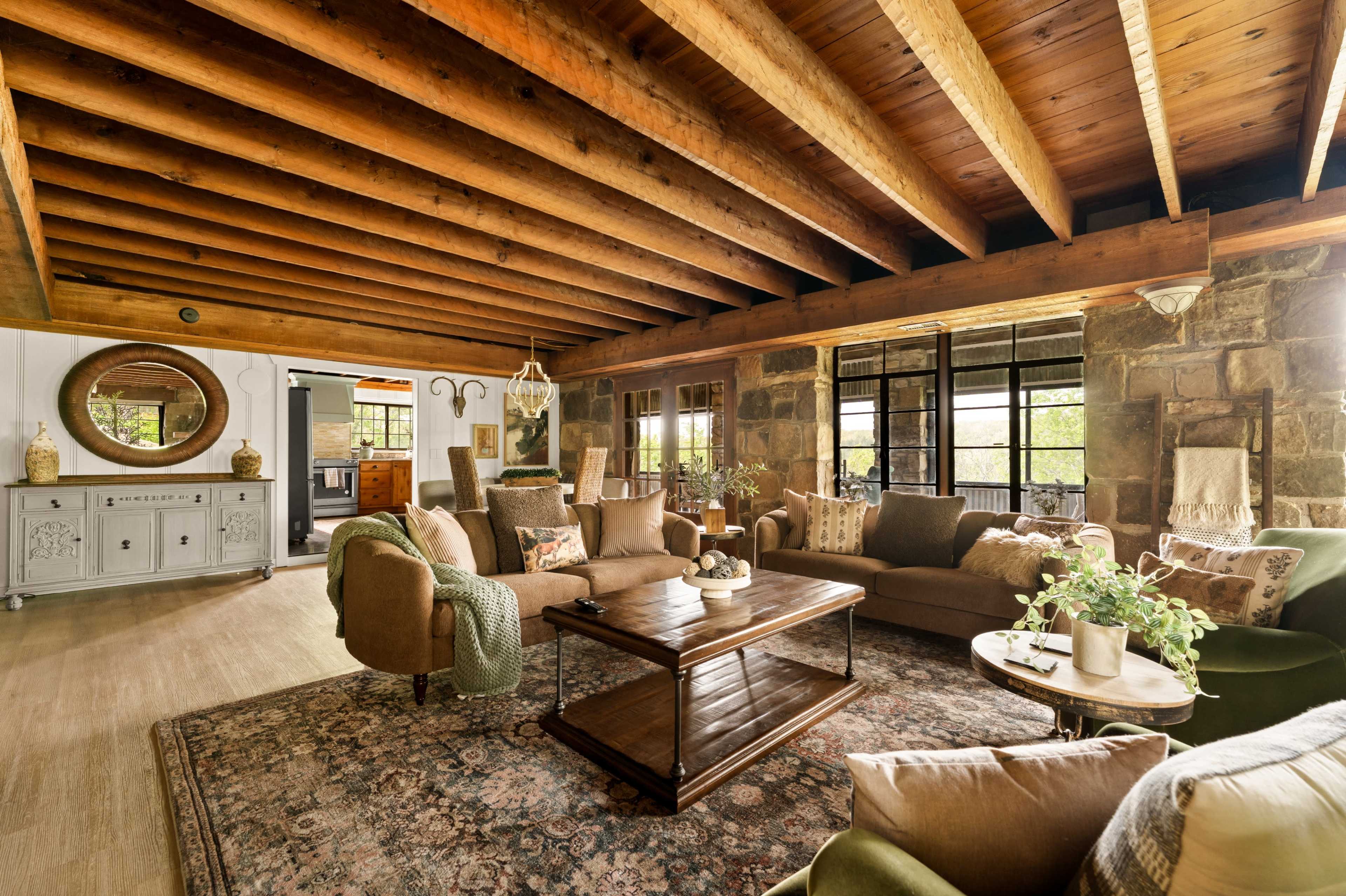The image shows a cozy living room with exposed wooden beams, stone walls, and a large rug, featuring comfortable seating and decorative plants.